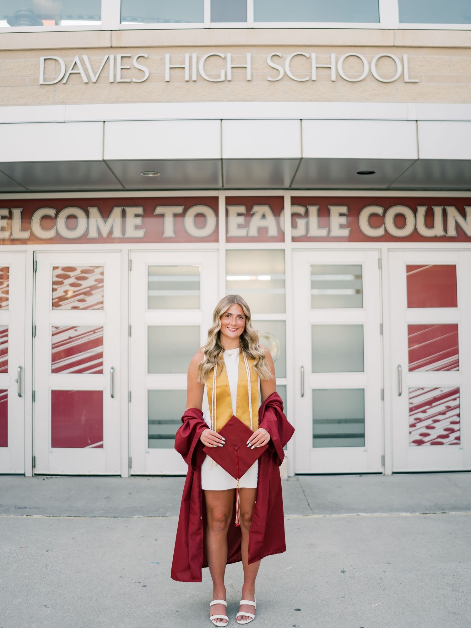 A smiling graduate poses in a maroon cap and gown outside Davies High School, holding her diploma in front of the entrance.