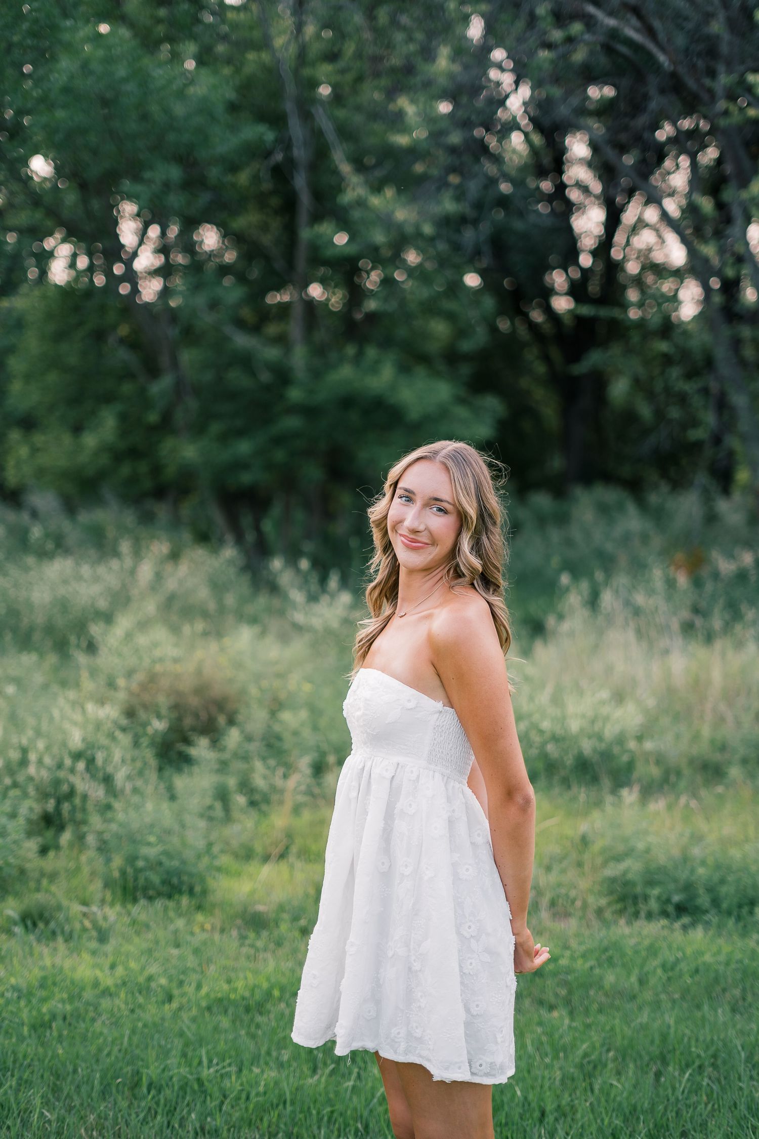 A person in a flowing white sundress poses playfully in a lush green outdoor setting during golden hour.