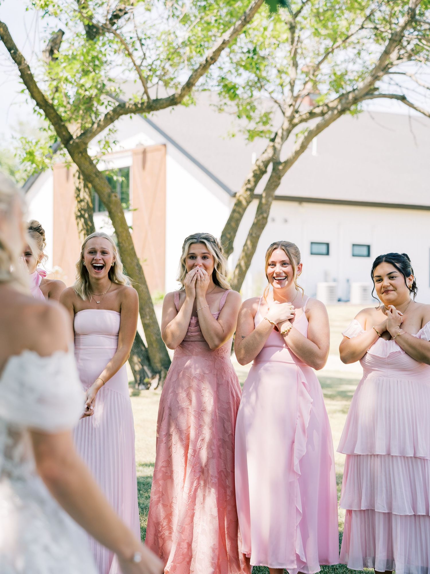 Bridesmaids in pink dresses react with joy and excitement seeing the bride for the first time at an outdoor wedding.
