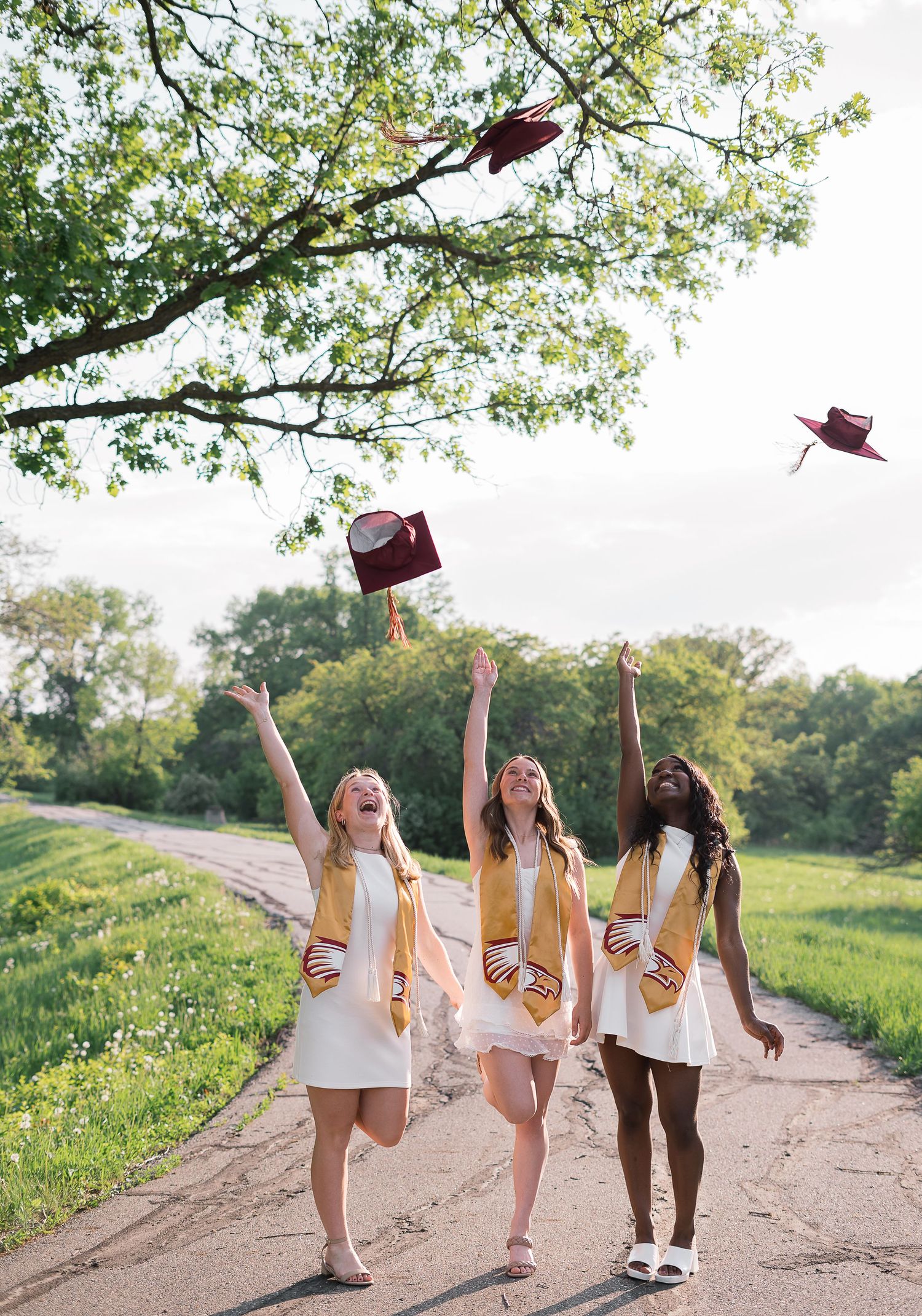 Three graduates in white dresses toss burgundy caps in the air while celebrating outdoors.