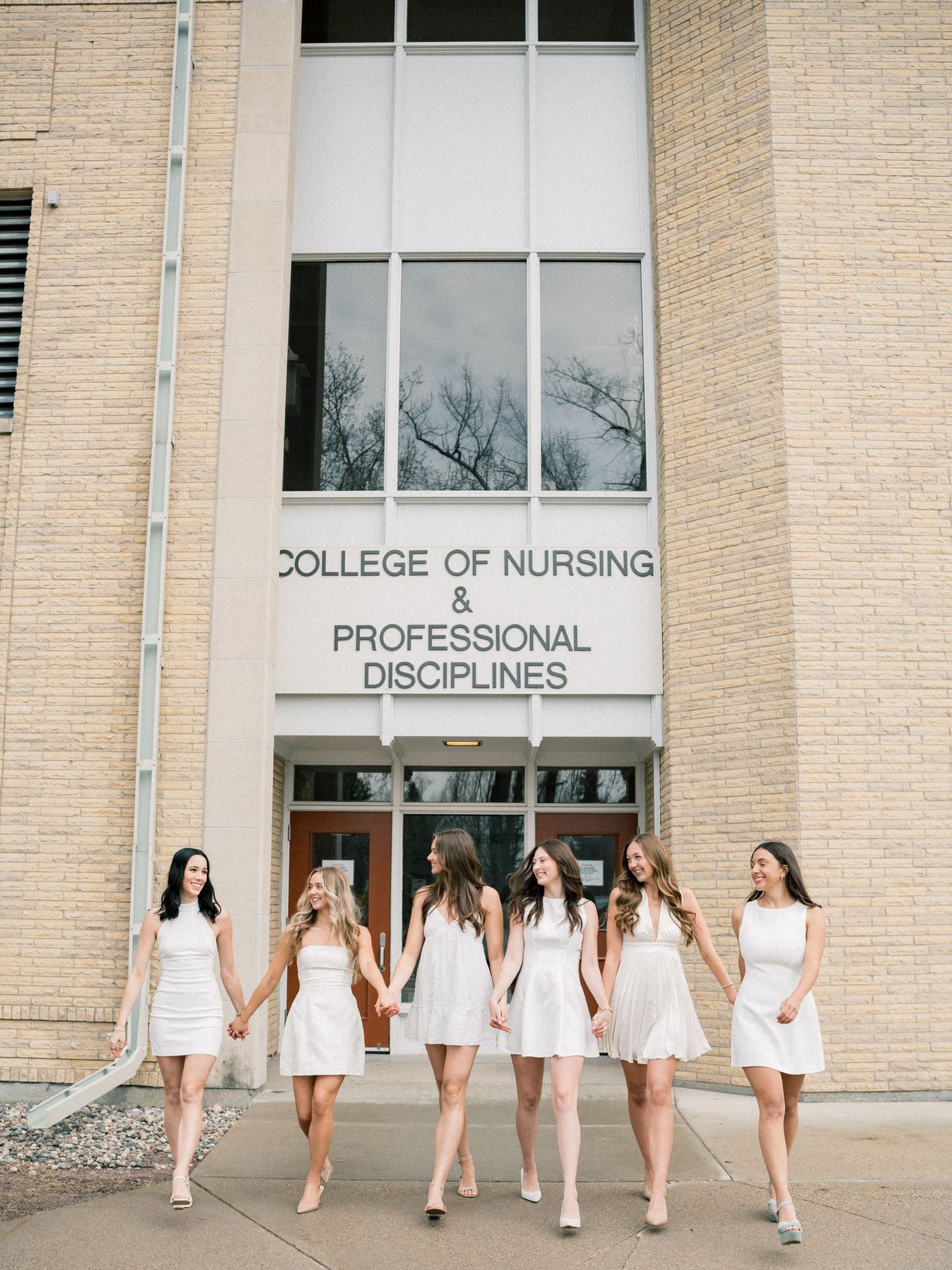 Six nursing graduates in white dresses hold hands walking in front of College of Nursing & Professional Disciplines building.