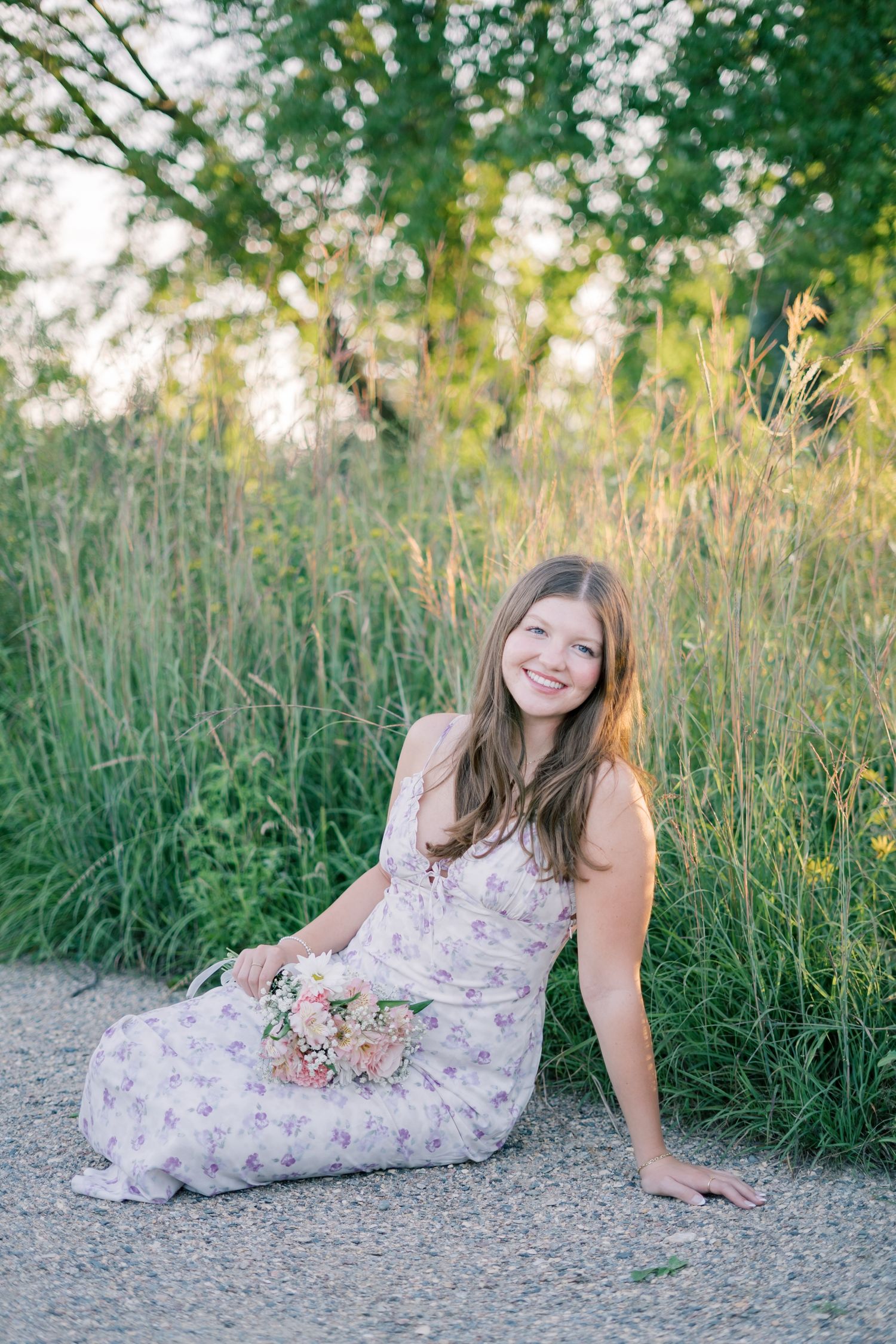Someone in a floral dress poses gracefully on a gravel path surrounded by tall summer grasses at golden hour.