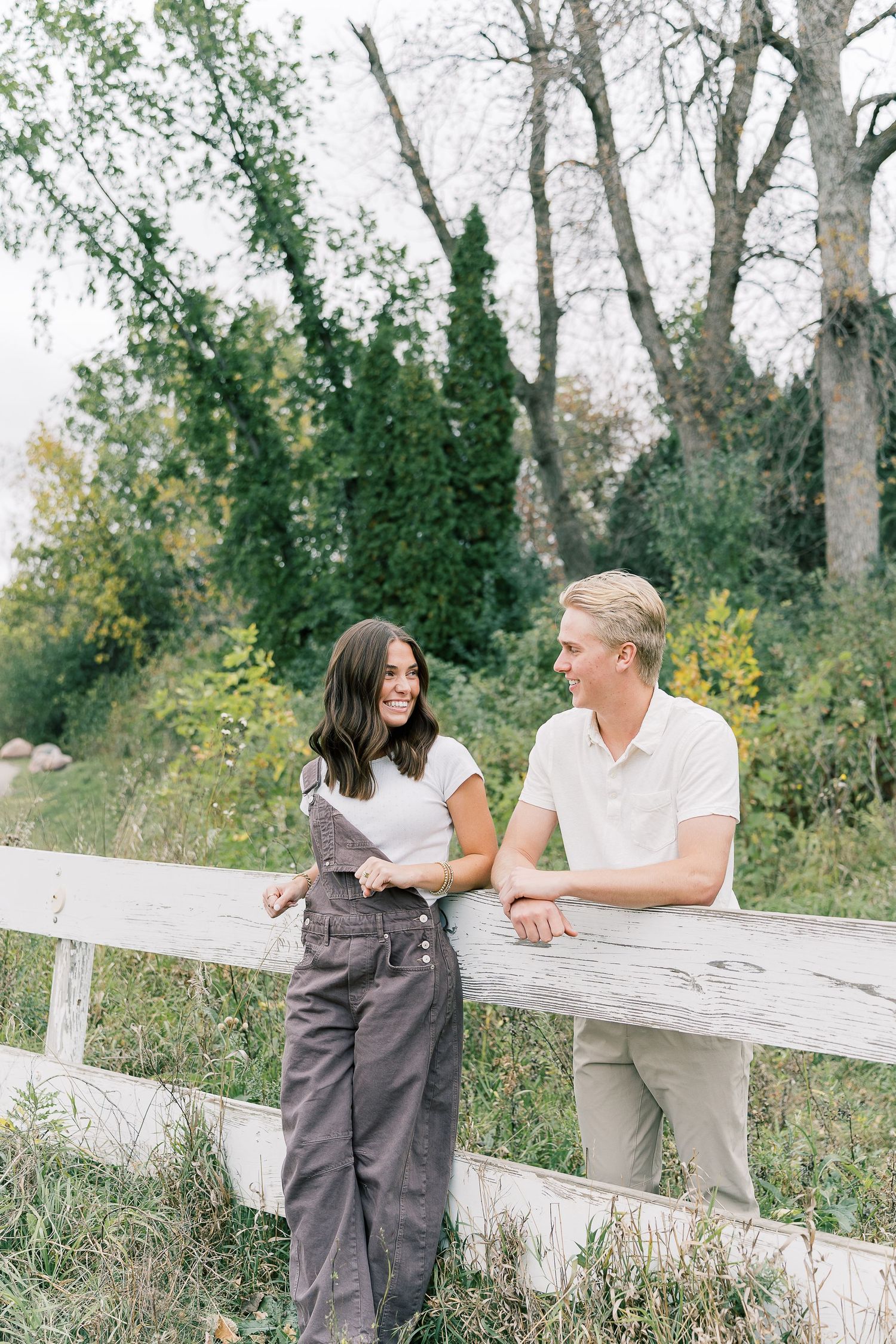A couple shares a tender moment leaning against a white fence in a scenic outdoor garden setting.