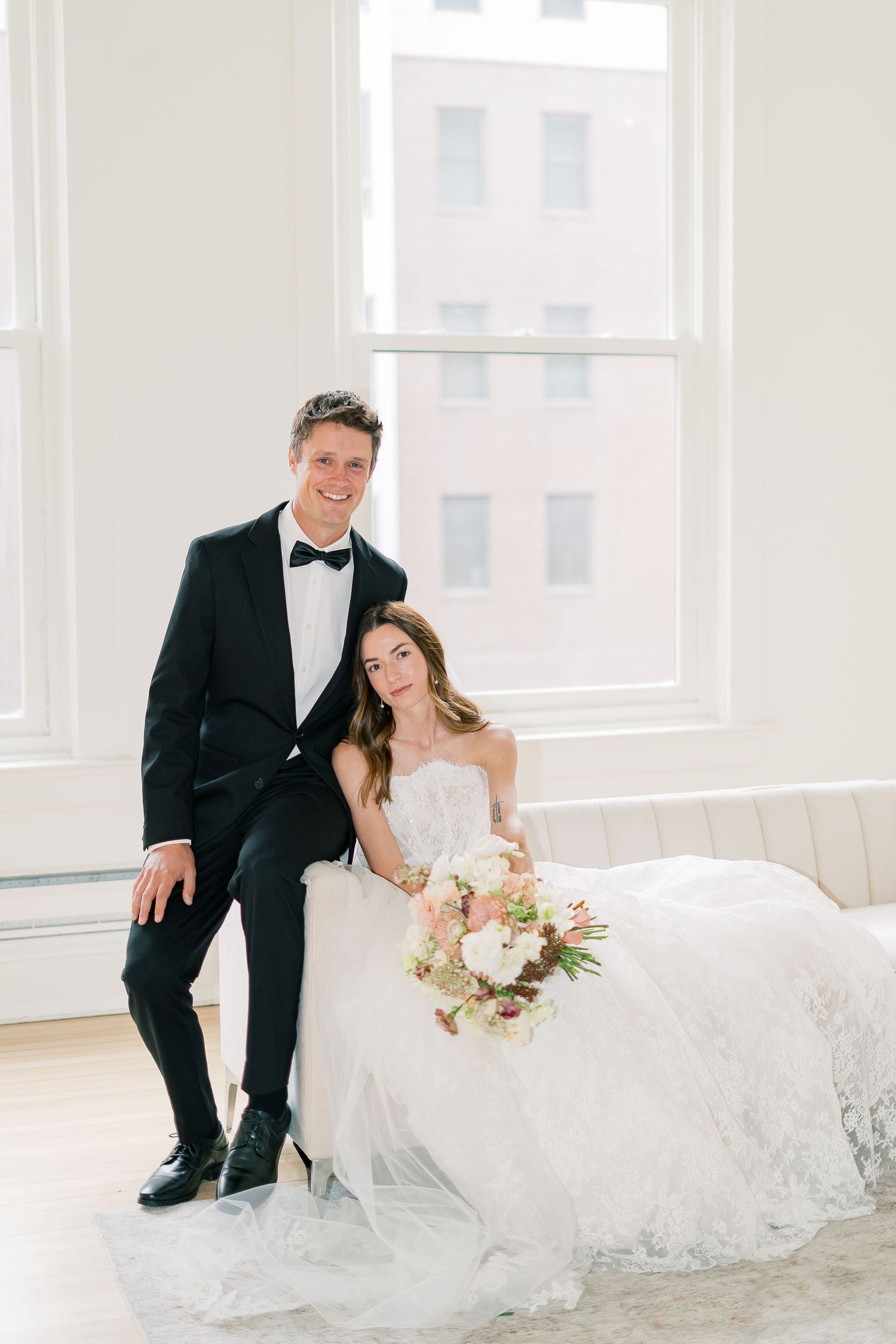A bride in a flowing white gown sits with her bouquet next to her groom in a classic black tuxedo by a bright window.