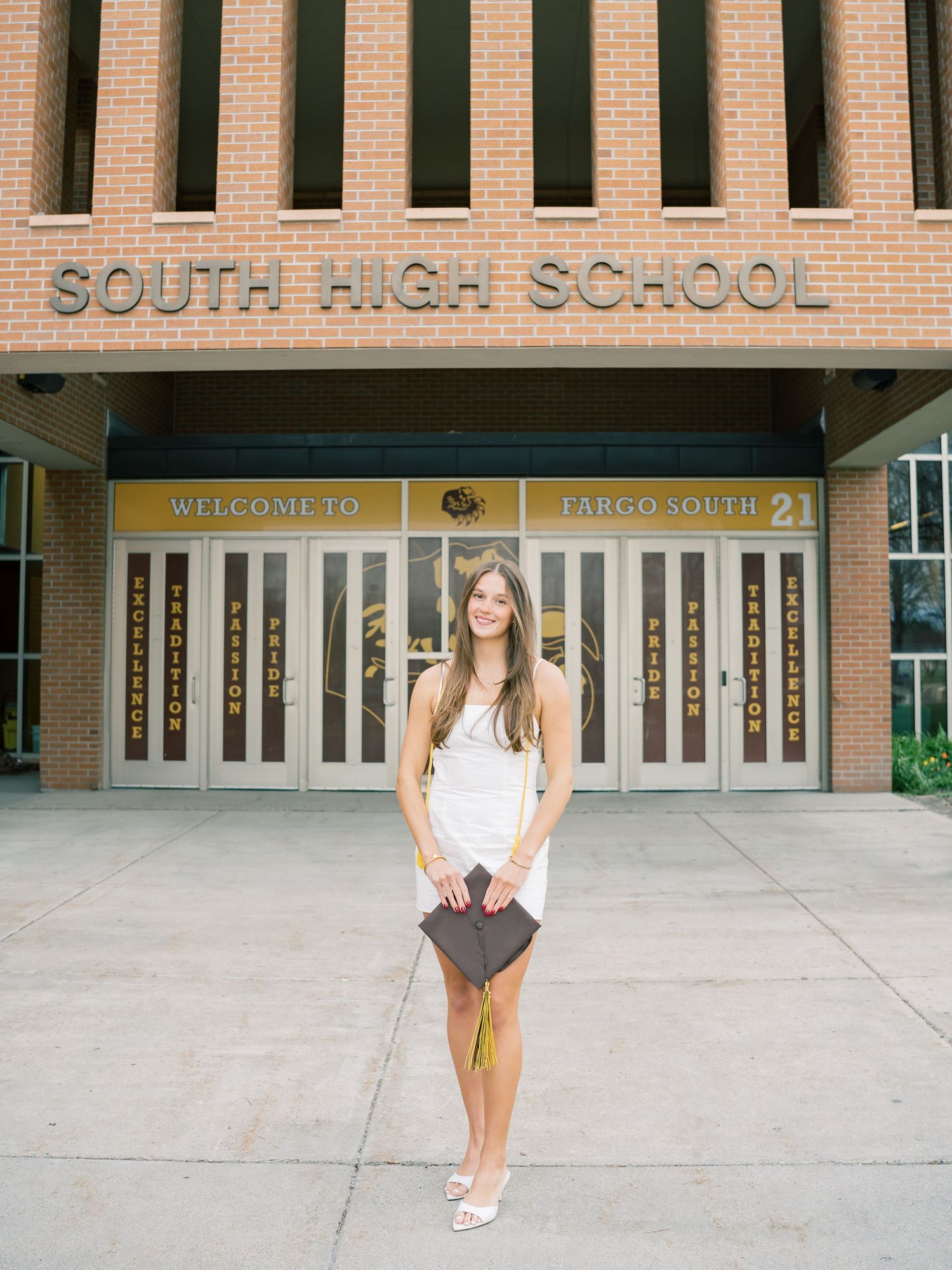 A young woman in a white dress holds a diploma folder outside Fargo South High School entrance.
