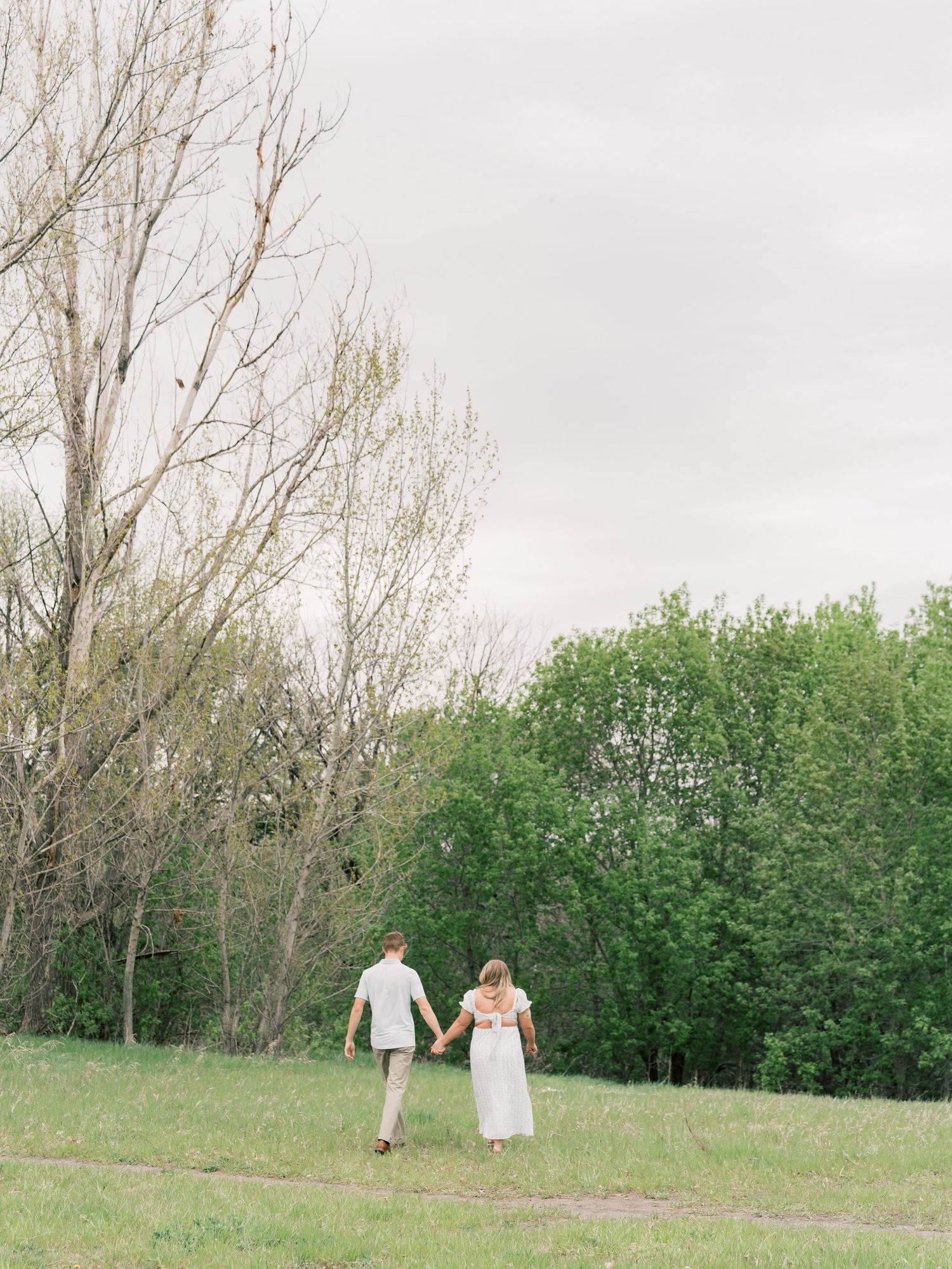 A couple in white attire walks hand-in-hand through a lush green meadow surrounded by trees under an overcast sky.