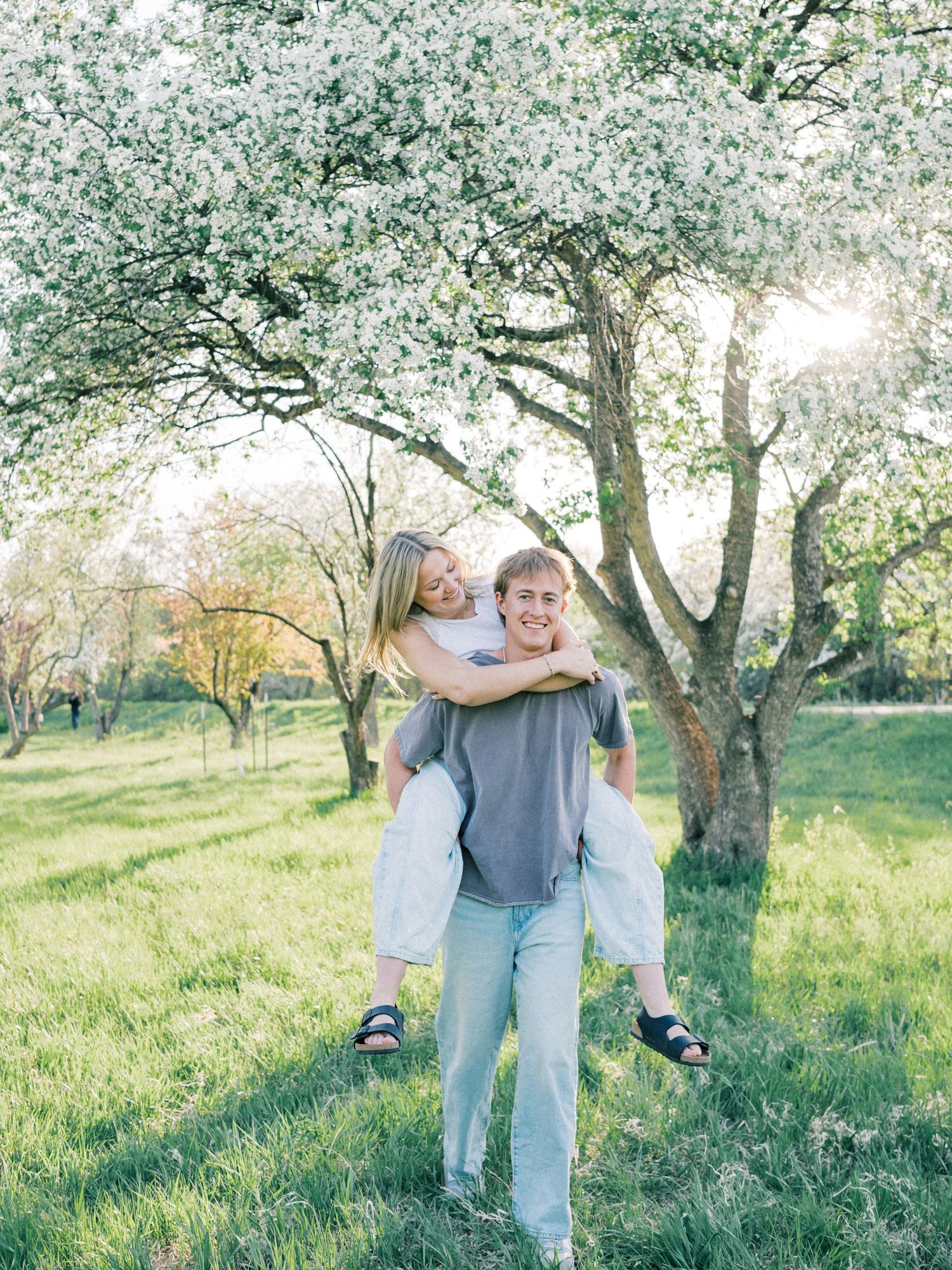 A couple shares a joyful piggyback ride under blooming white trees in a lush green meadow during spring.