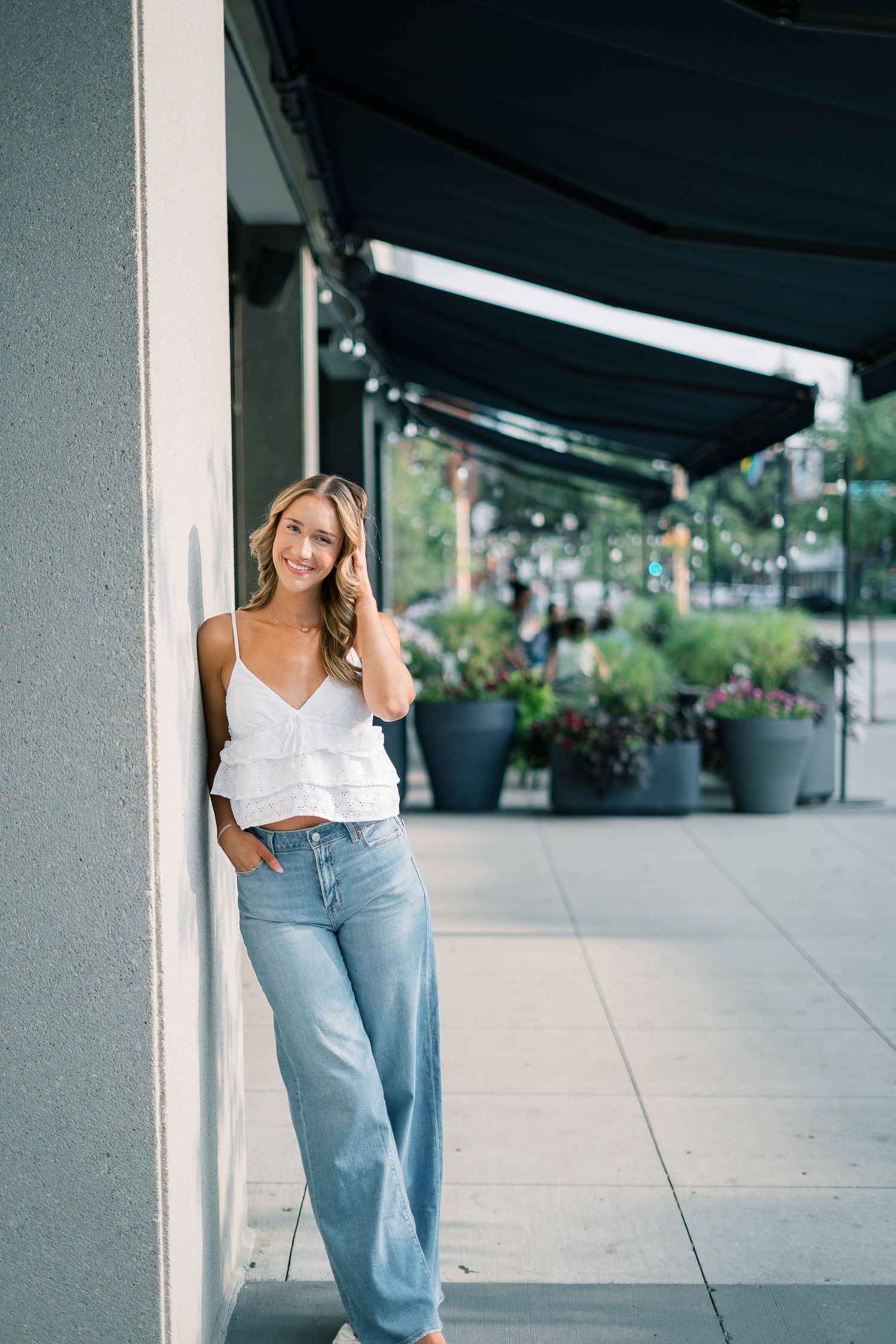 Woman in white top and light blue jeans poses casually against outdoor column in urban setting.