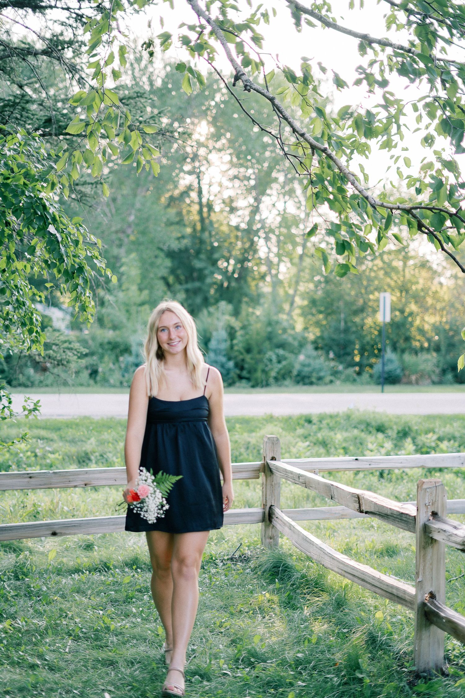 A person in a black summer dress stands along a rustic wooden fence surrounded by lush greenery on a sunny day.