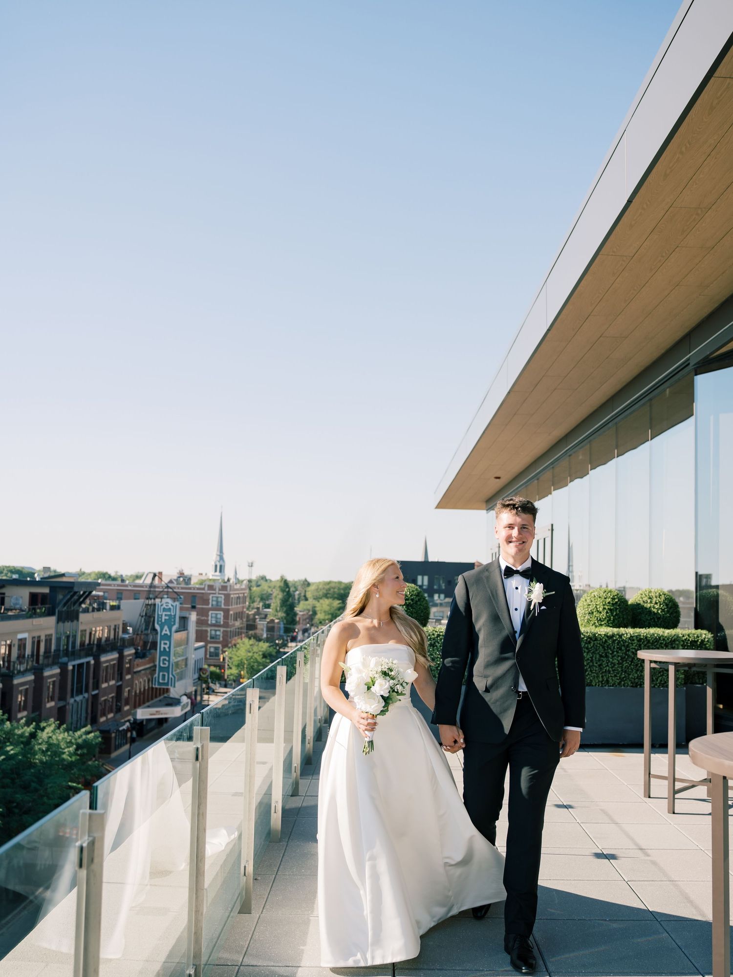 Bride and groom walk hand-in-hand on a rooftop terrace, with city skyline views on a sunny wedding day.