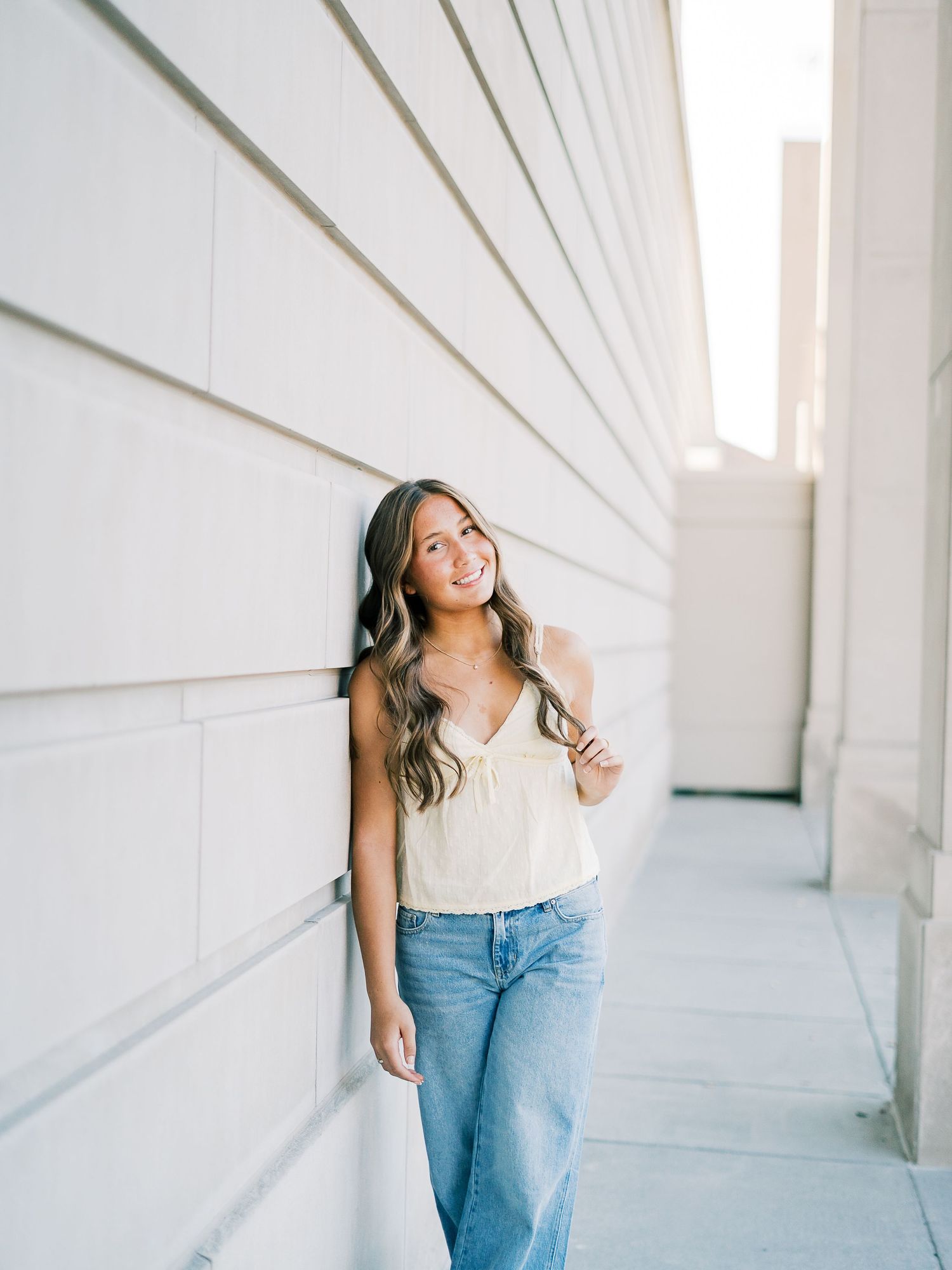 A person wearing a white top and light blue jeans poses casually against a white wall in a bright hallway.
