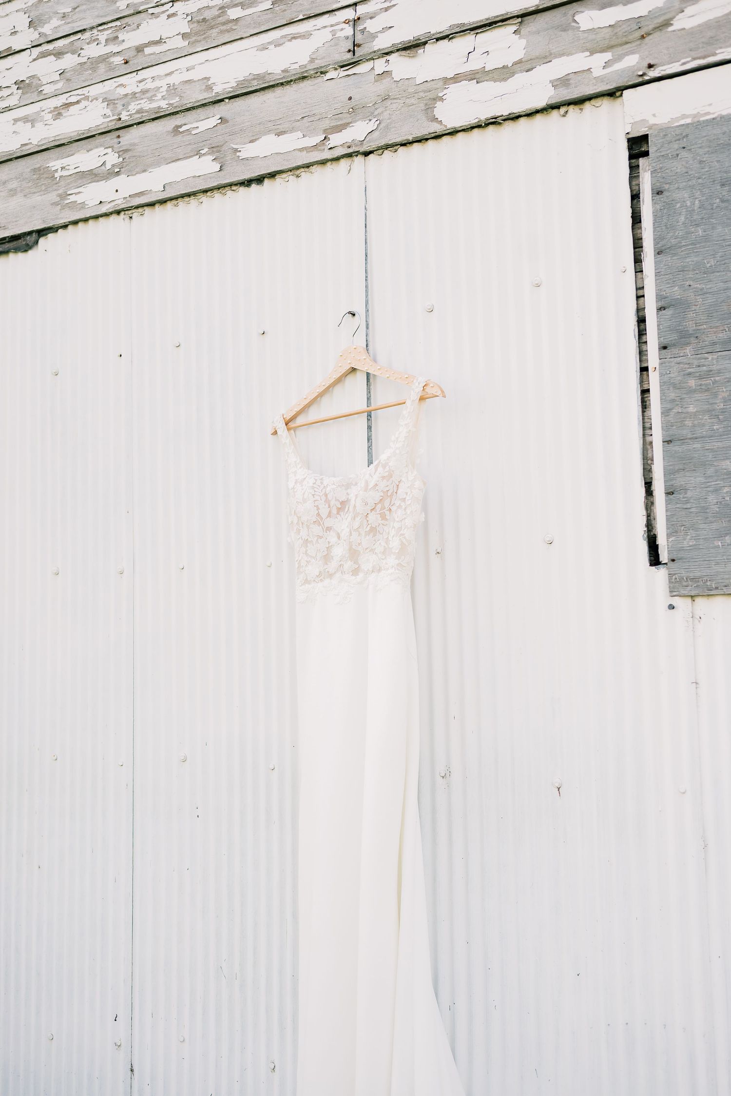White wedding dress hanging against a rustic wooden wall with natural lighting creating an ethereal glow.