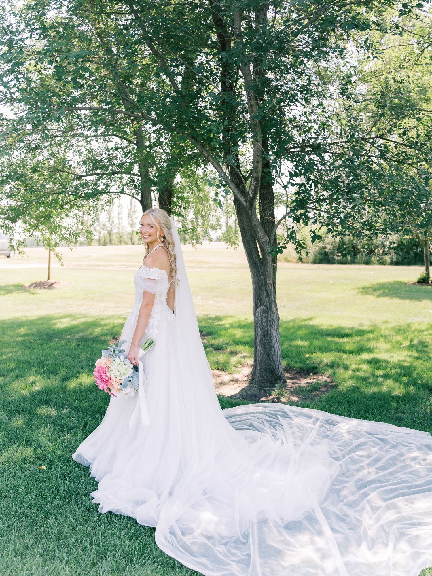 Bride in off-shoulder white gown with long veil stands on lush green lawn beside trees holding colorful bouquet.
