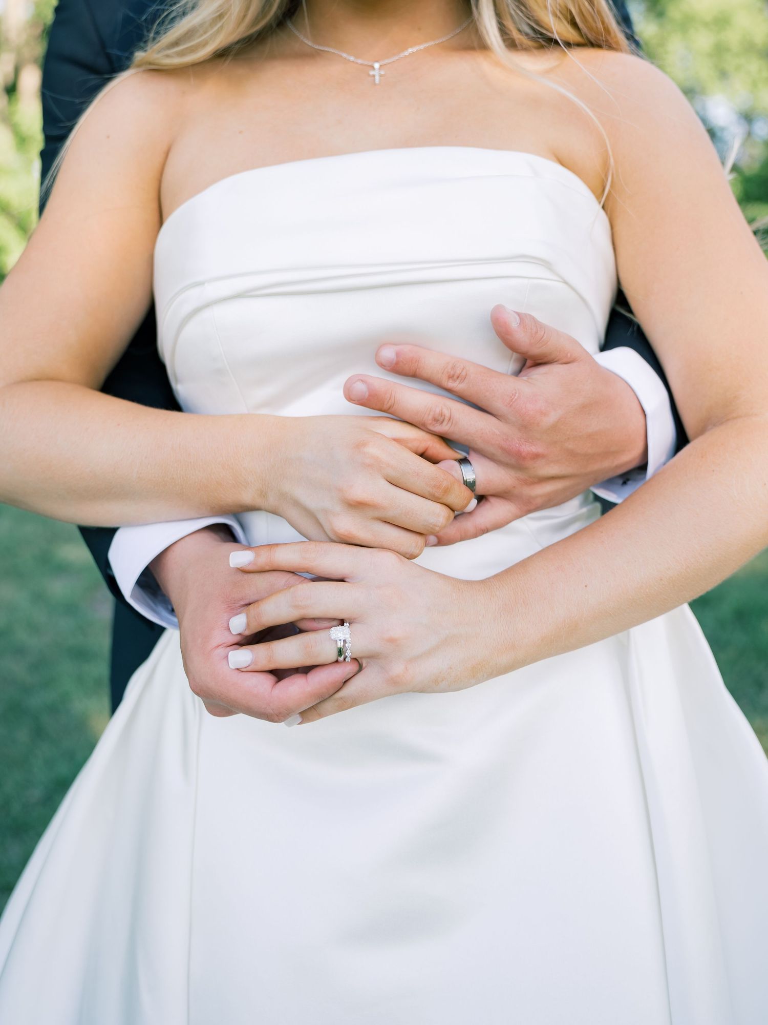 Close-up of newlyweds hands showing wedding rings, bride in strapless white gown embraced by groom in dark suit.