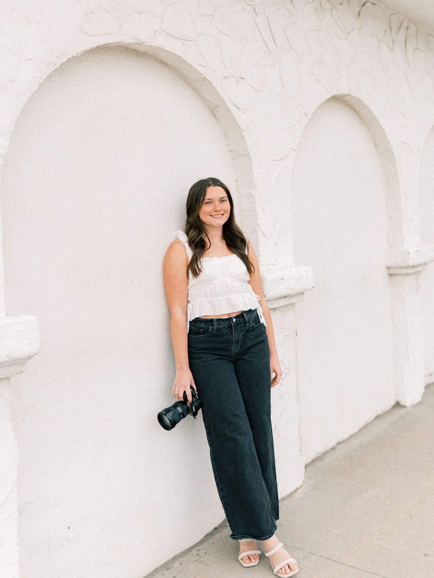 Young woman in white crop top and dark jeans holding a camera, posing against white arched wall.