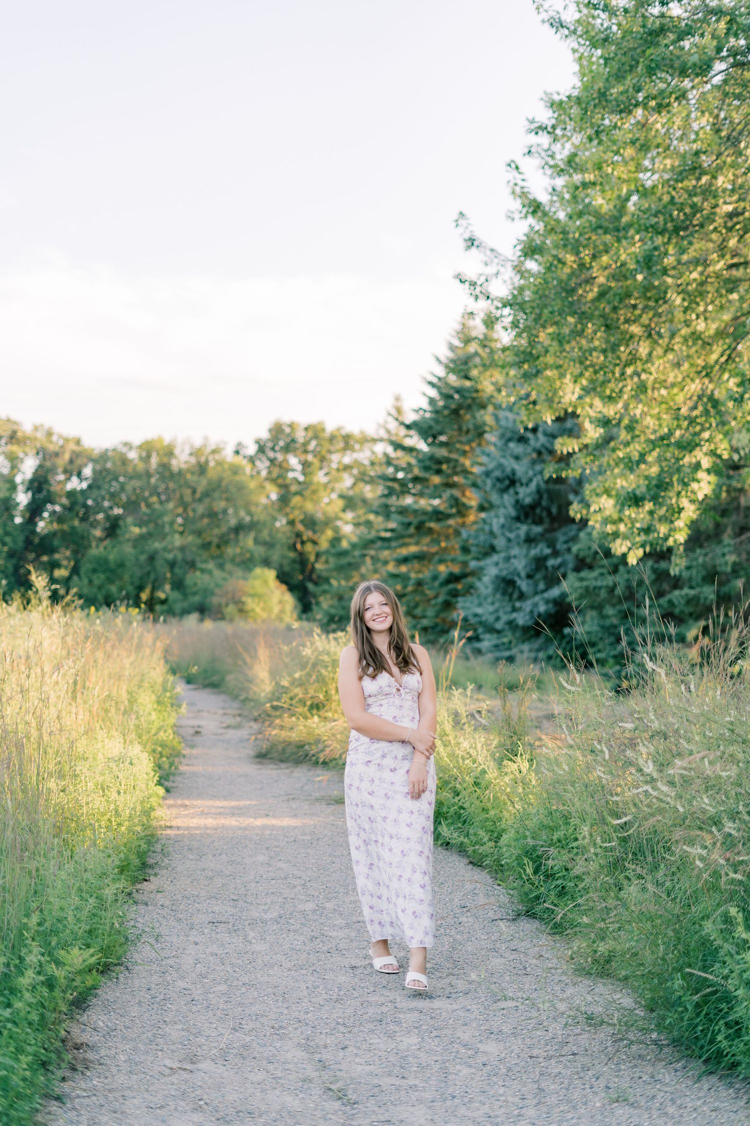 A person in a white floral dress walks along a scenic nature trail bordered by green foliage and trees in summer light.