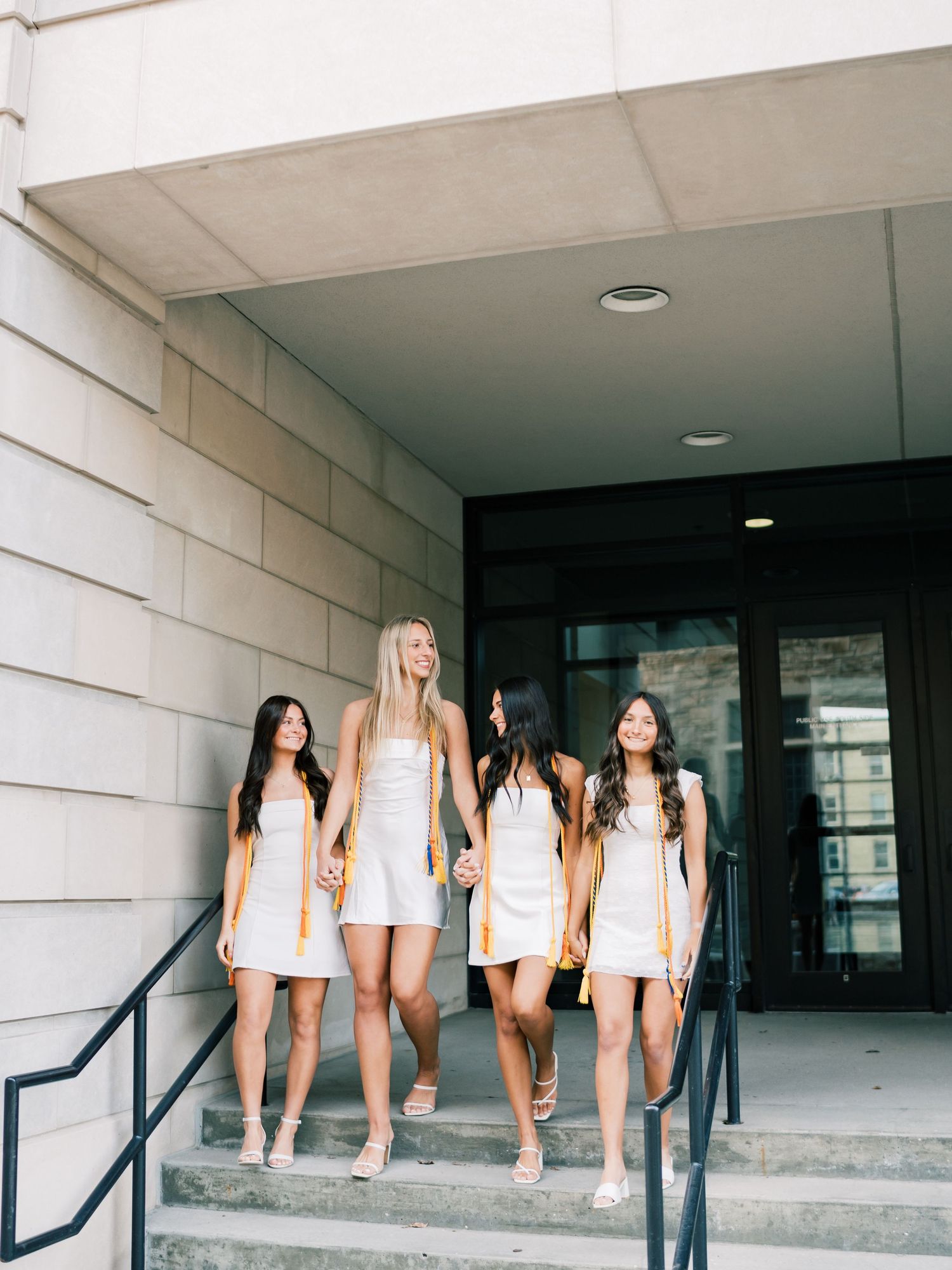 Four young women in white dresses and gold graduation stoles walk down steps outside a university building.