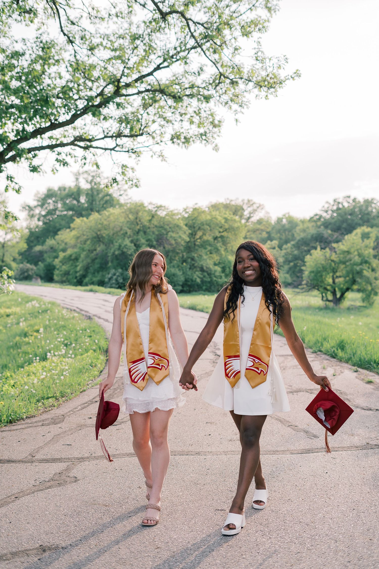 Two graduates in white dresses and pink stoles walk together holding graduation caps on a scenic path.
