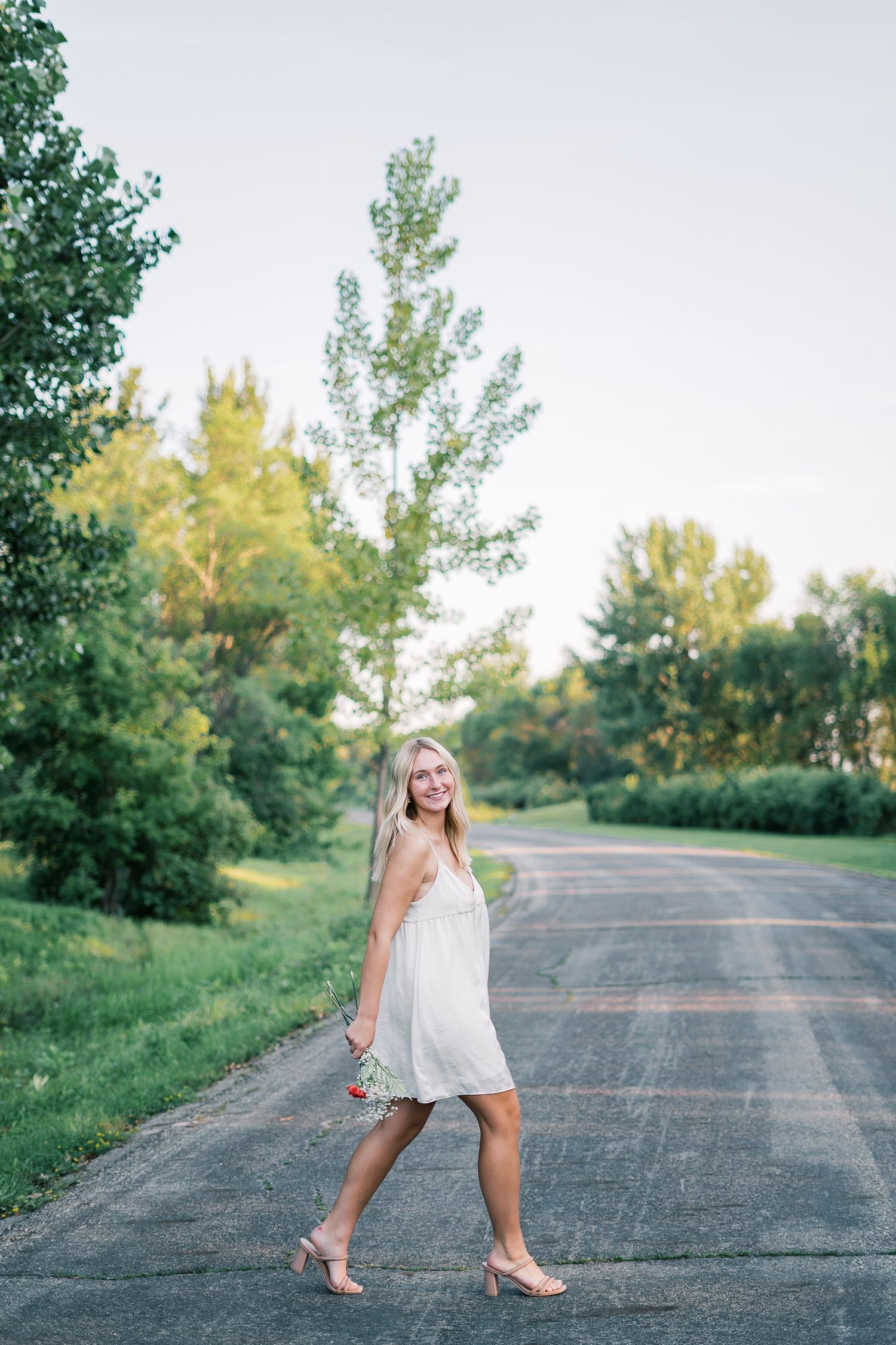 A person in a white dress twirls playfully on a rural road surrounded by green trees on a sunny summer day.