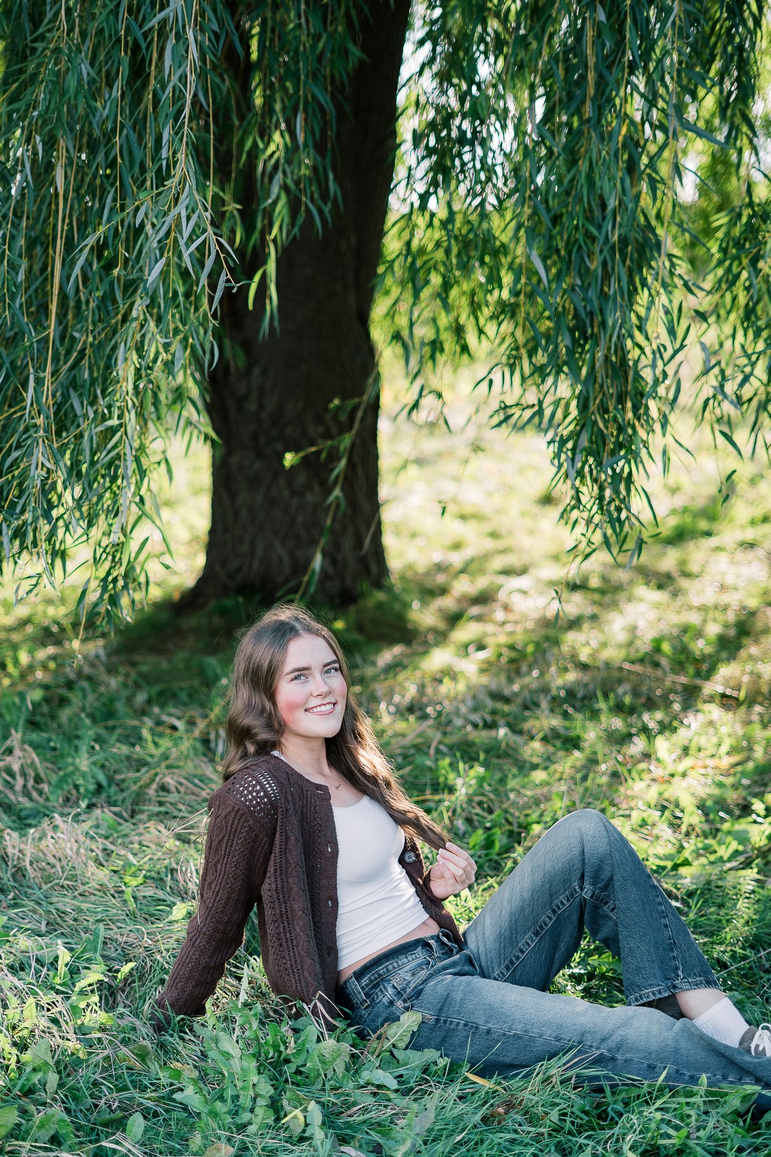 A person relaxes under a weeping willow tree on a sunny day wearing jeans and a brown cardigan.