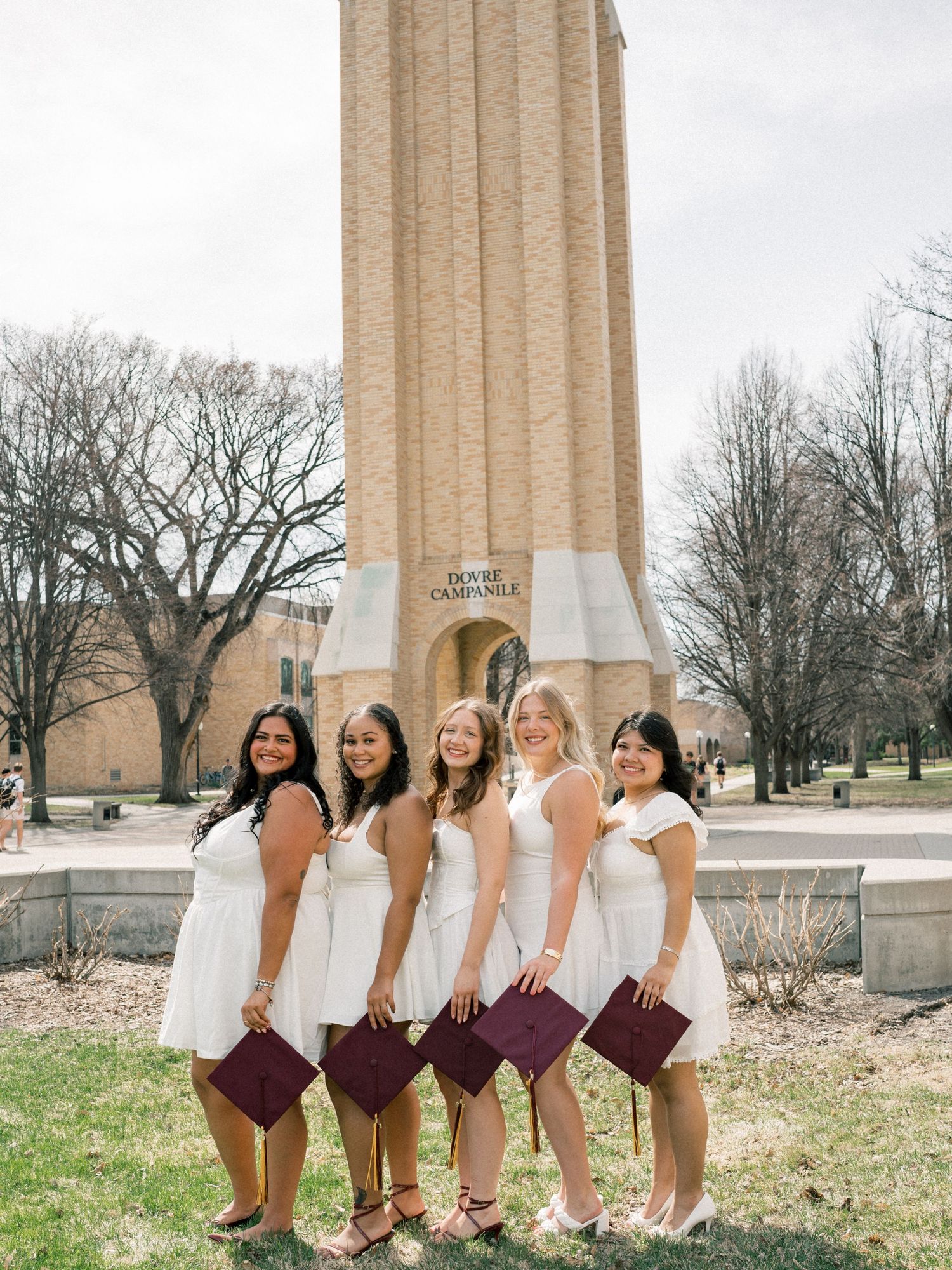 Five graduates in white dresses hold maroon caps in front of Doud Campanile on a college campus.