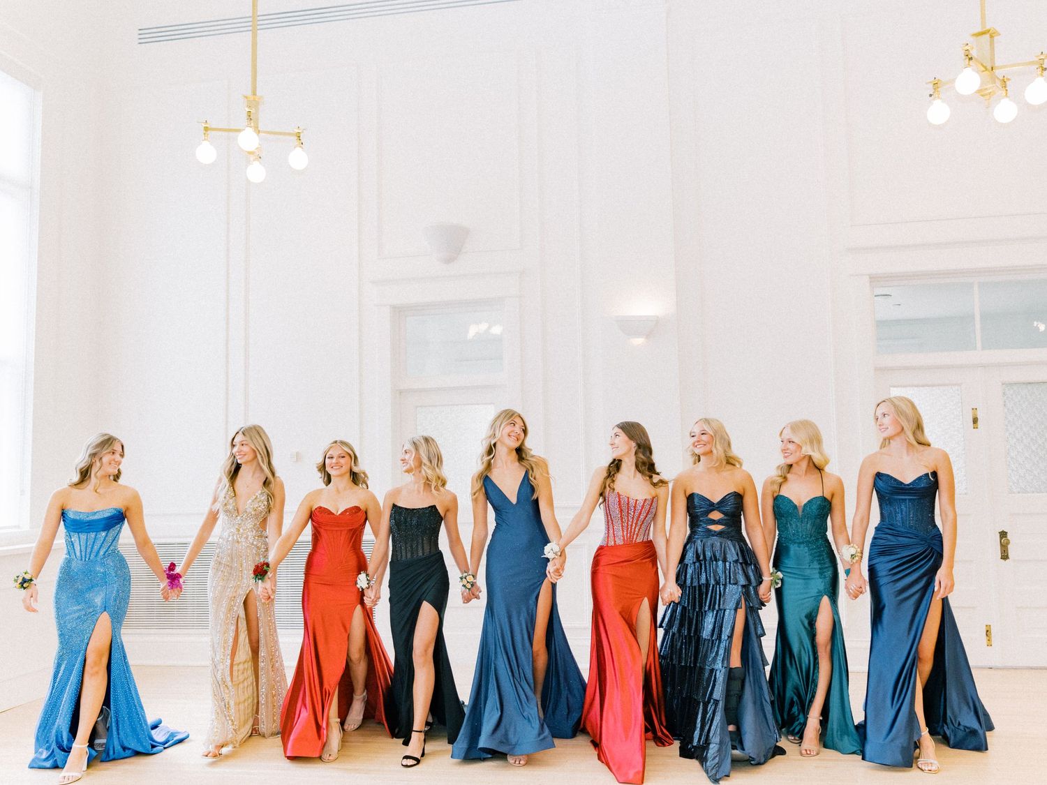 Nine bridesmaids in colorful satin gowns walk together in a bright white ballroom, showcasing mismatched dress styles.