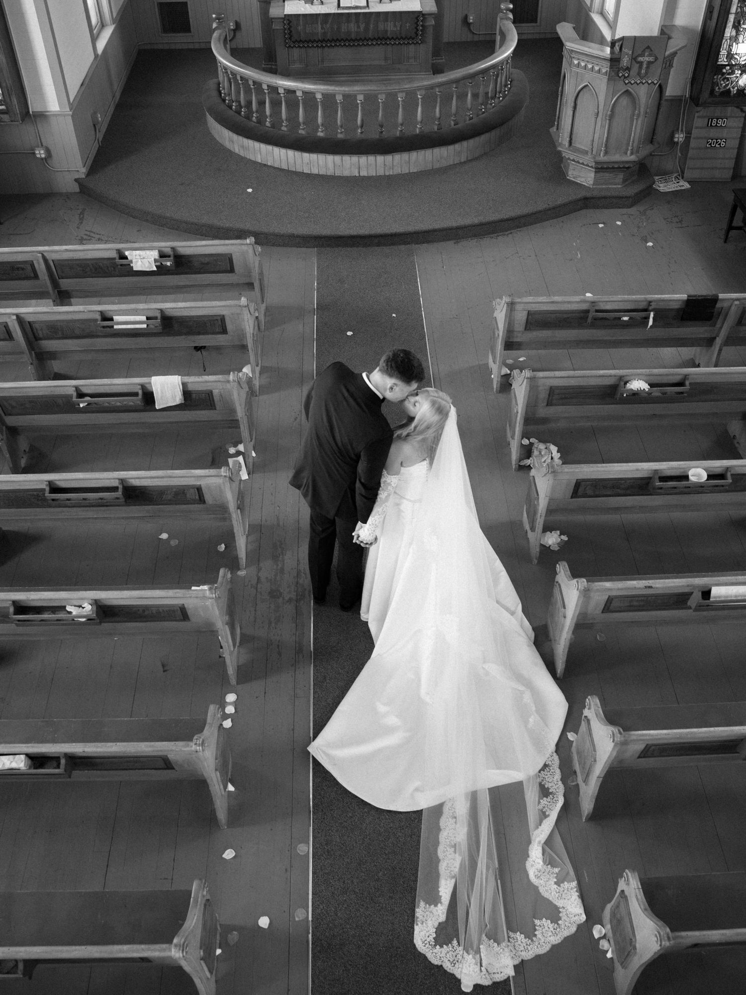Aerial black and white photo of bride and groom sharing a kiss in church aisle, wedding gown train spread elegantly.