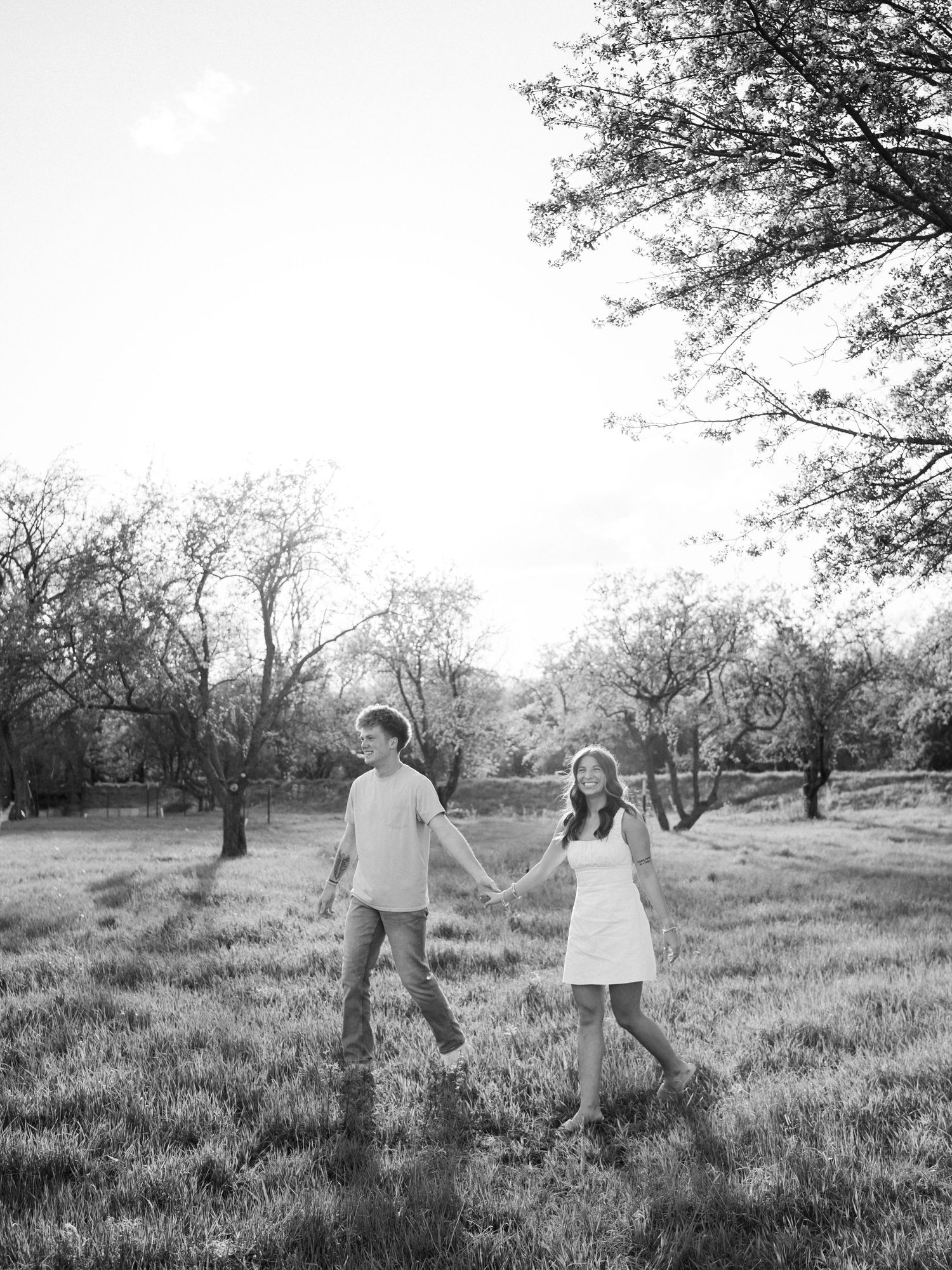 Black and white photo of a couple holding hands while walking through a sunlit field surrounded by trees.