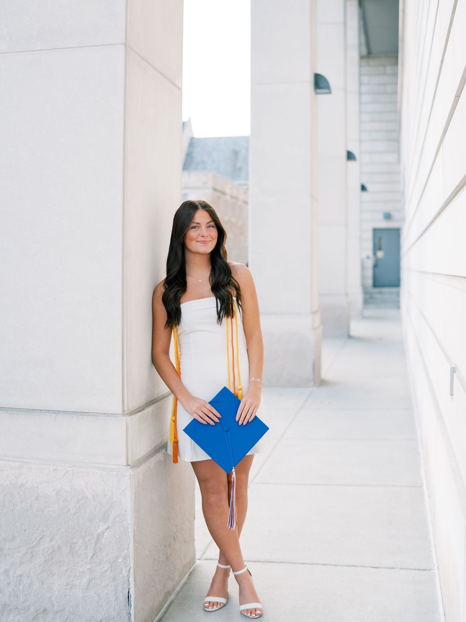 A young woman in a white dress holds a blue graduation cap and gold stole, leaning against a column outdoors.