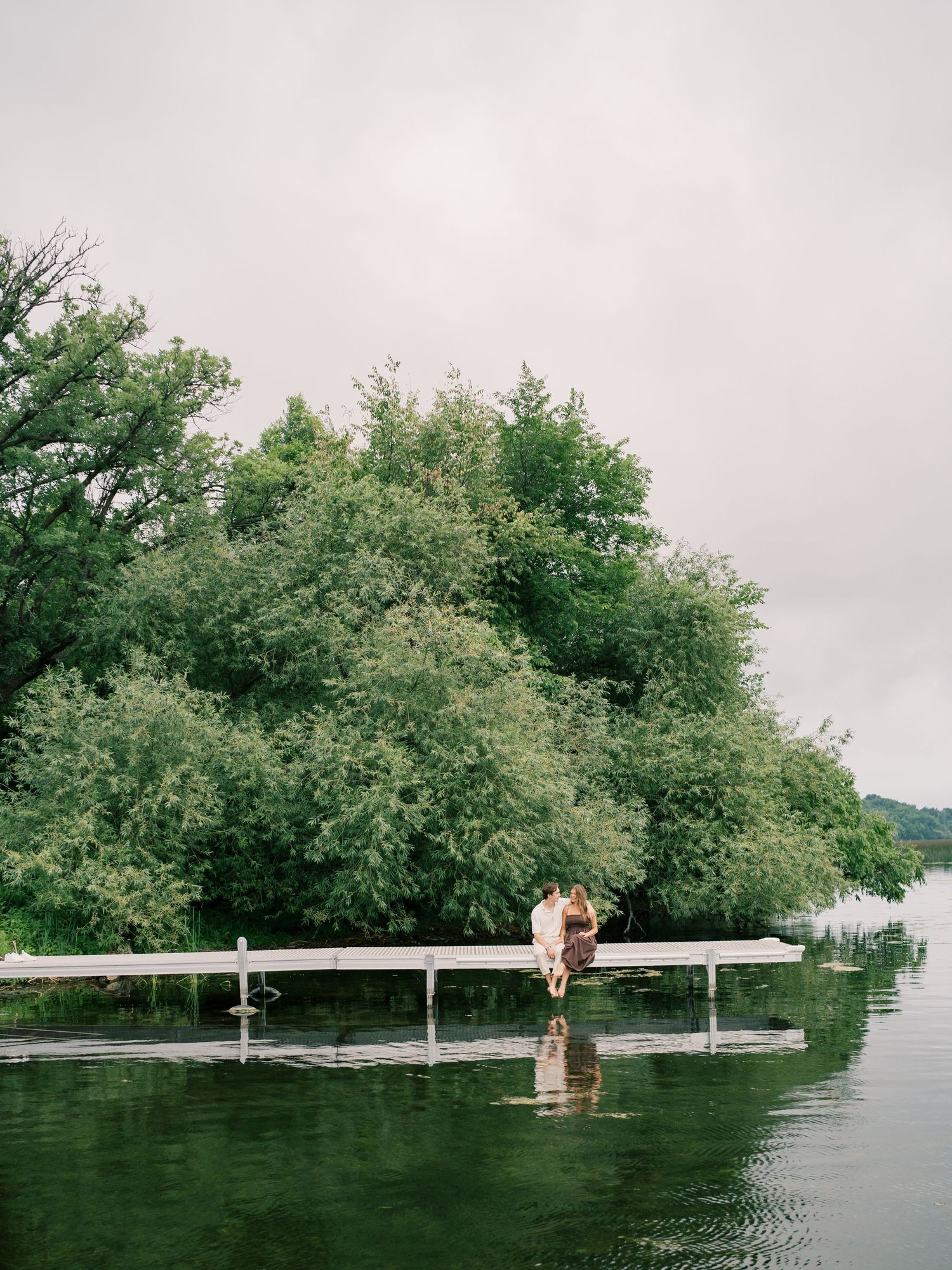 Couple sitting together on a wooden dock over calm green lake water surrounded by lush trees on a cloudy day.