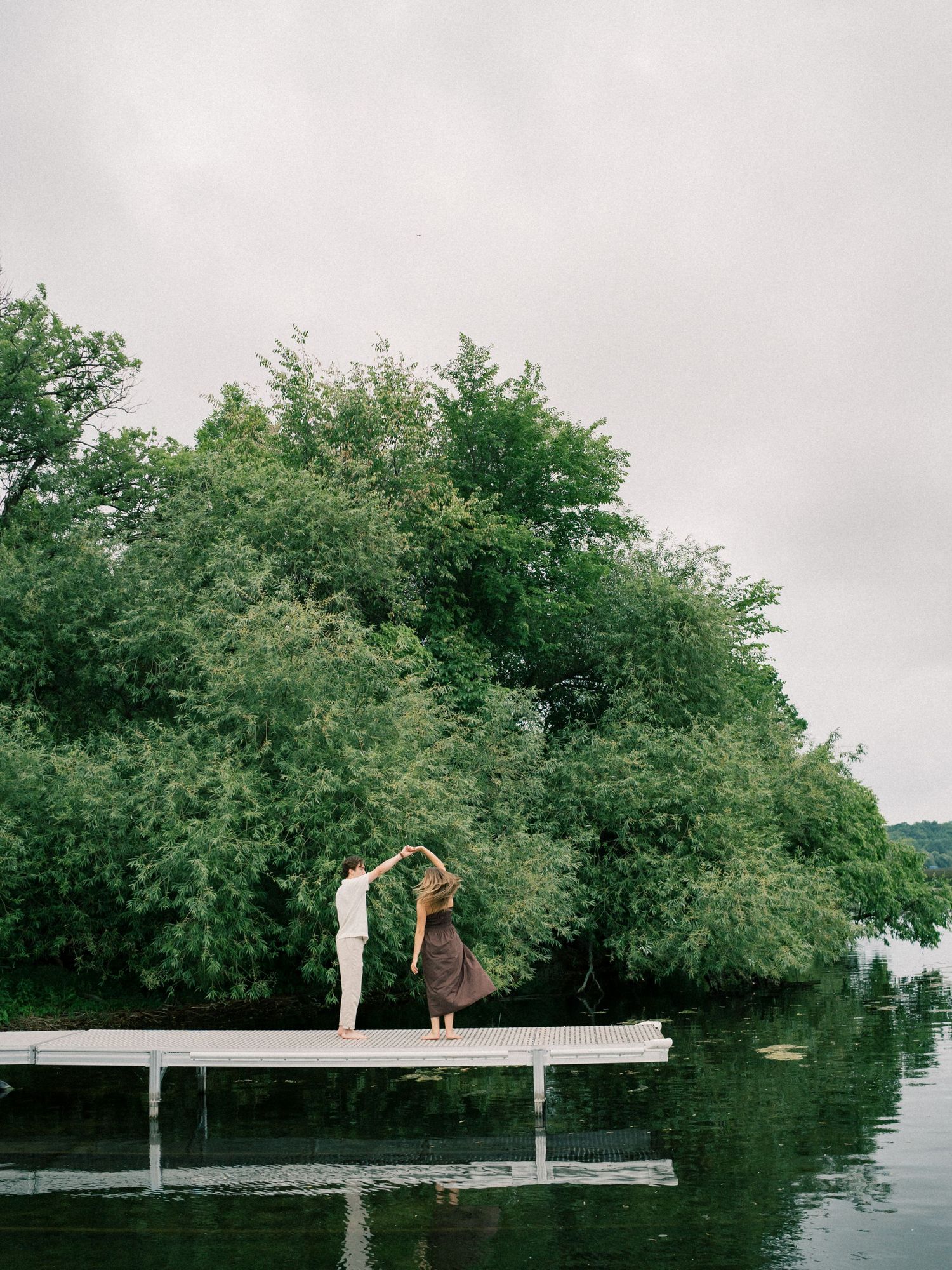 Couple dancing on a wooden dock surrounded by lush green trees and calm lake waters on an overcast day.