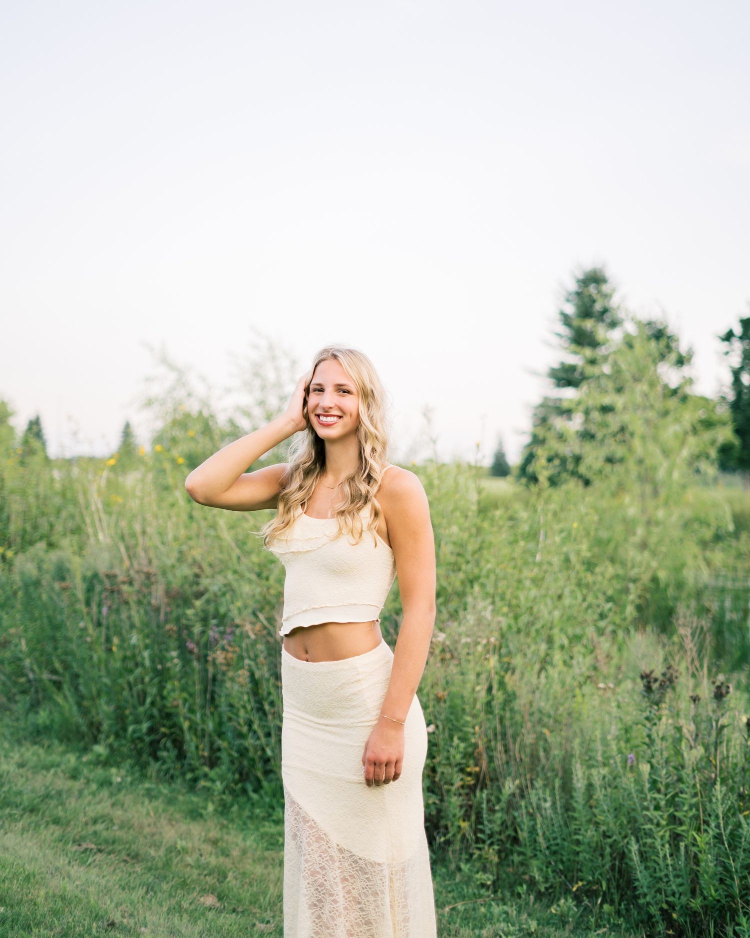 A person in a white lace dress poses gracefully in a field of tall grass and wildflowers at sunset.