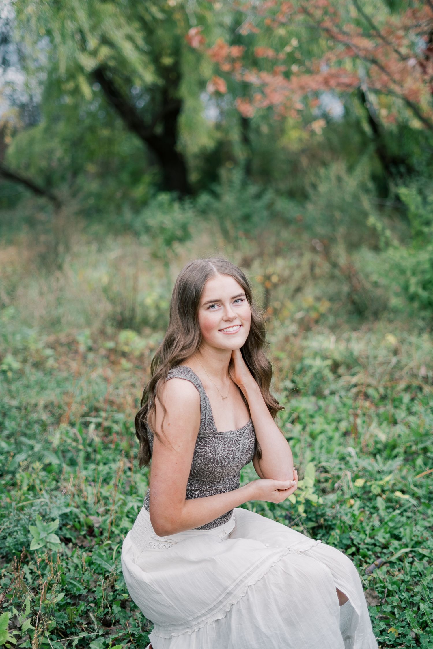 A person in a gray sleeveless top and white skirt sits in a lush garden setting with autumn foliage in the background.