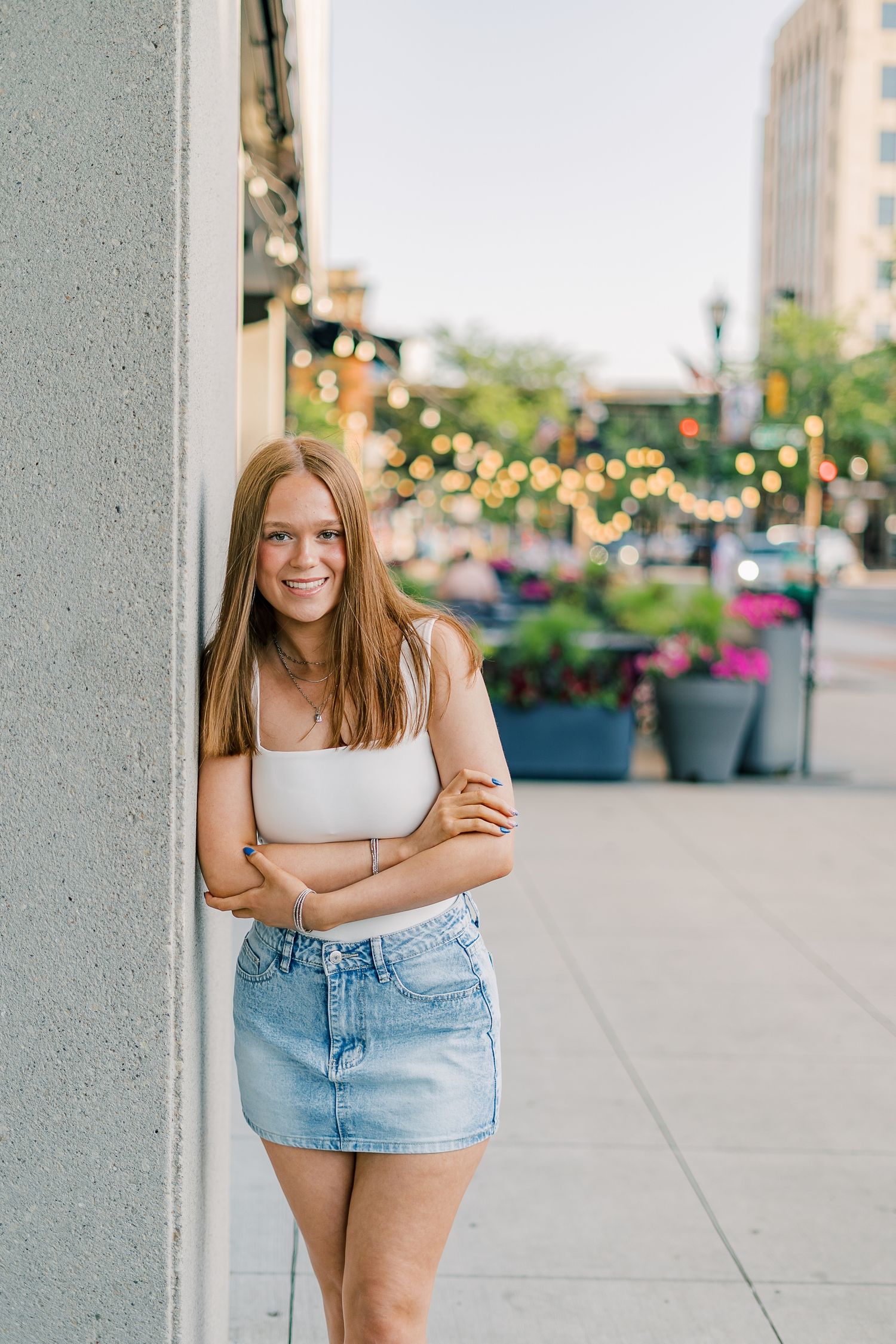 A person in a white tank top and denim shorts poses against a light gray column on a city sidewalk with planters.