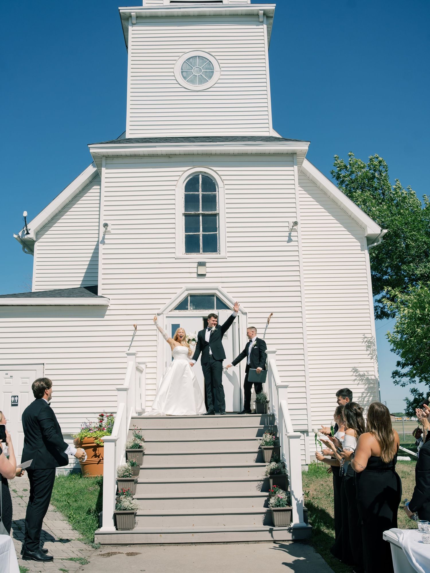 Joyful newlyweds celebrate on church steps in front of a white wooden chapel, surrounded by wedding guests on a sunny day.