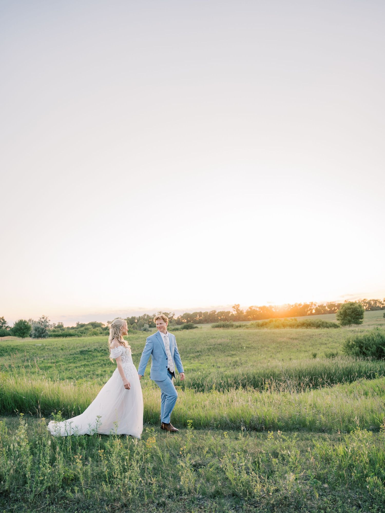 A bride and groom walk hand in hand through a lush green field during a golden sunset wedding photo.