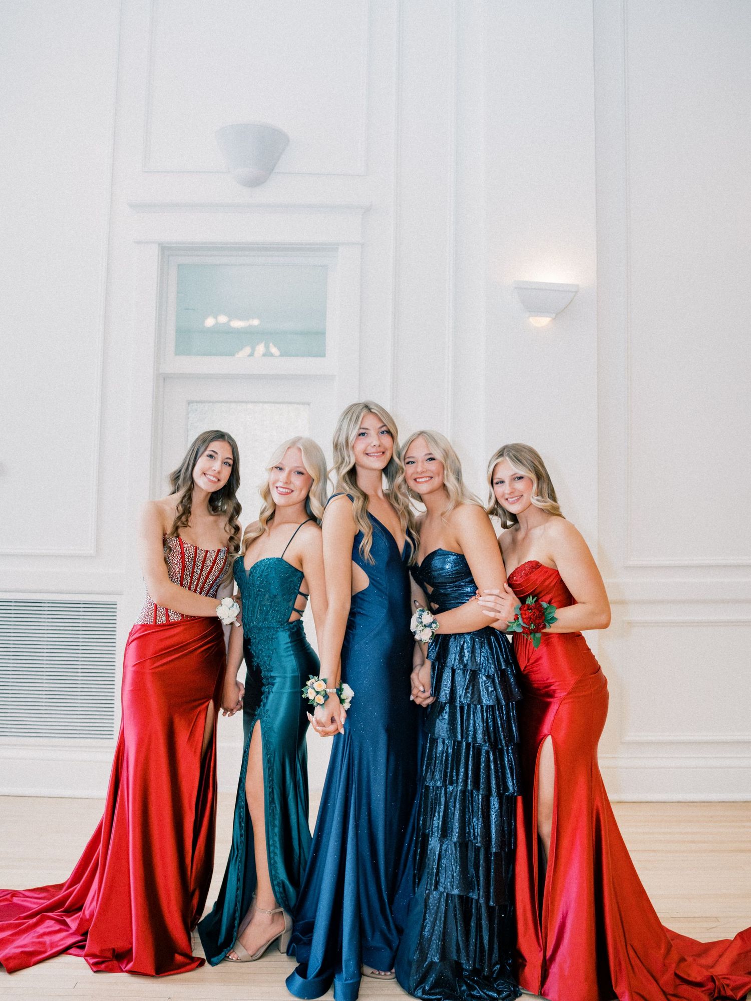 Five women in elegant formal gowns in red and navy blue pose together smiling at a prom or formal event.