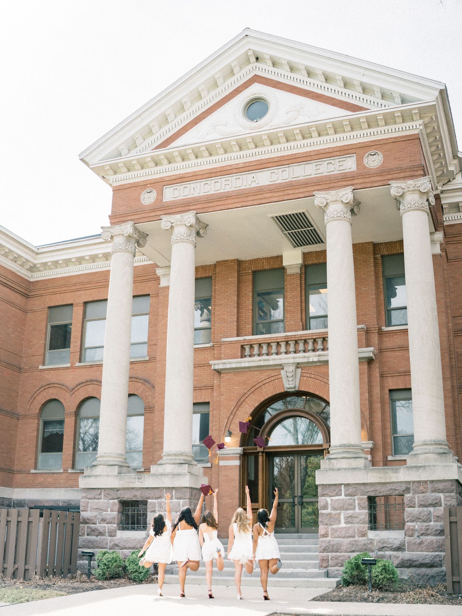 Bridesmaids in white dresses celebrate outside a brick college building with grand columns, tossing bouquets in the air.