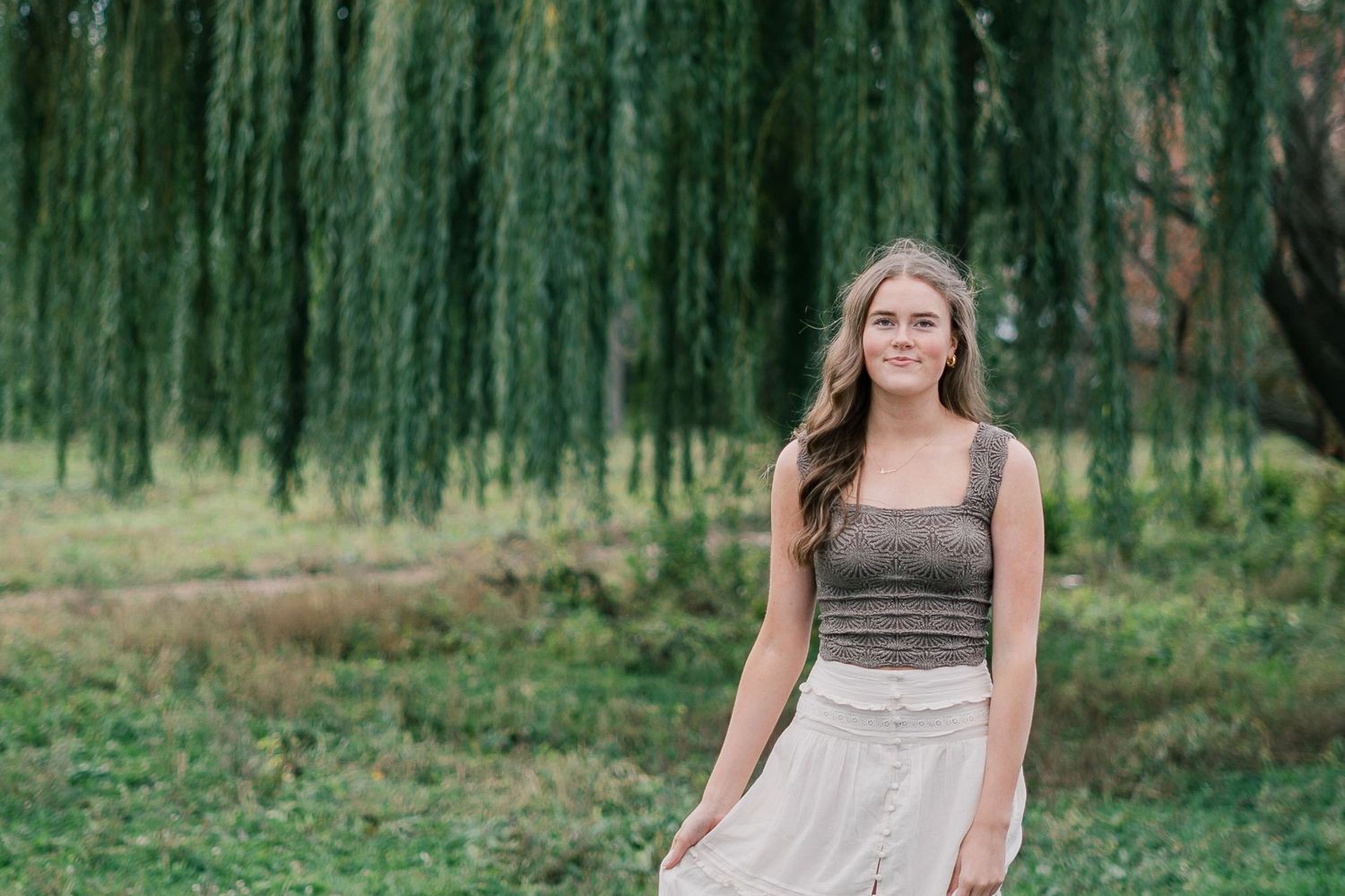 A woman in a brown and cream dress poses outdoors in a garden setting with willow trees in the background.