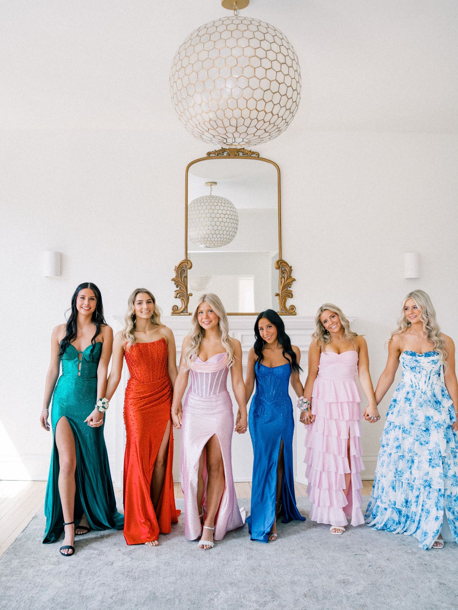 Six women in colorful formal gowns pose together holding hands in an elegant white room with a gold mirror.