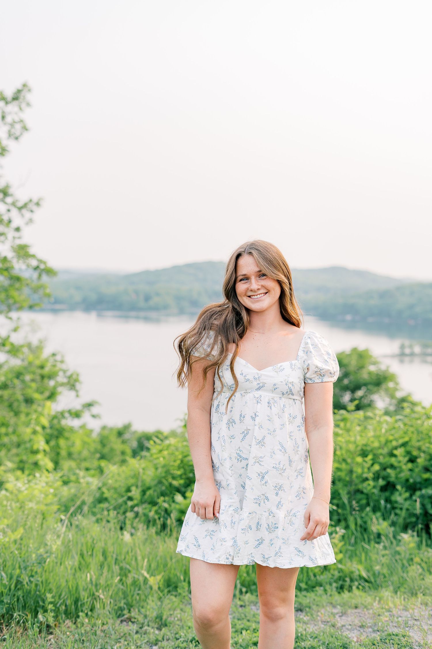 Woman in floral dress poses on hilltop overlooking a serene lake.