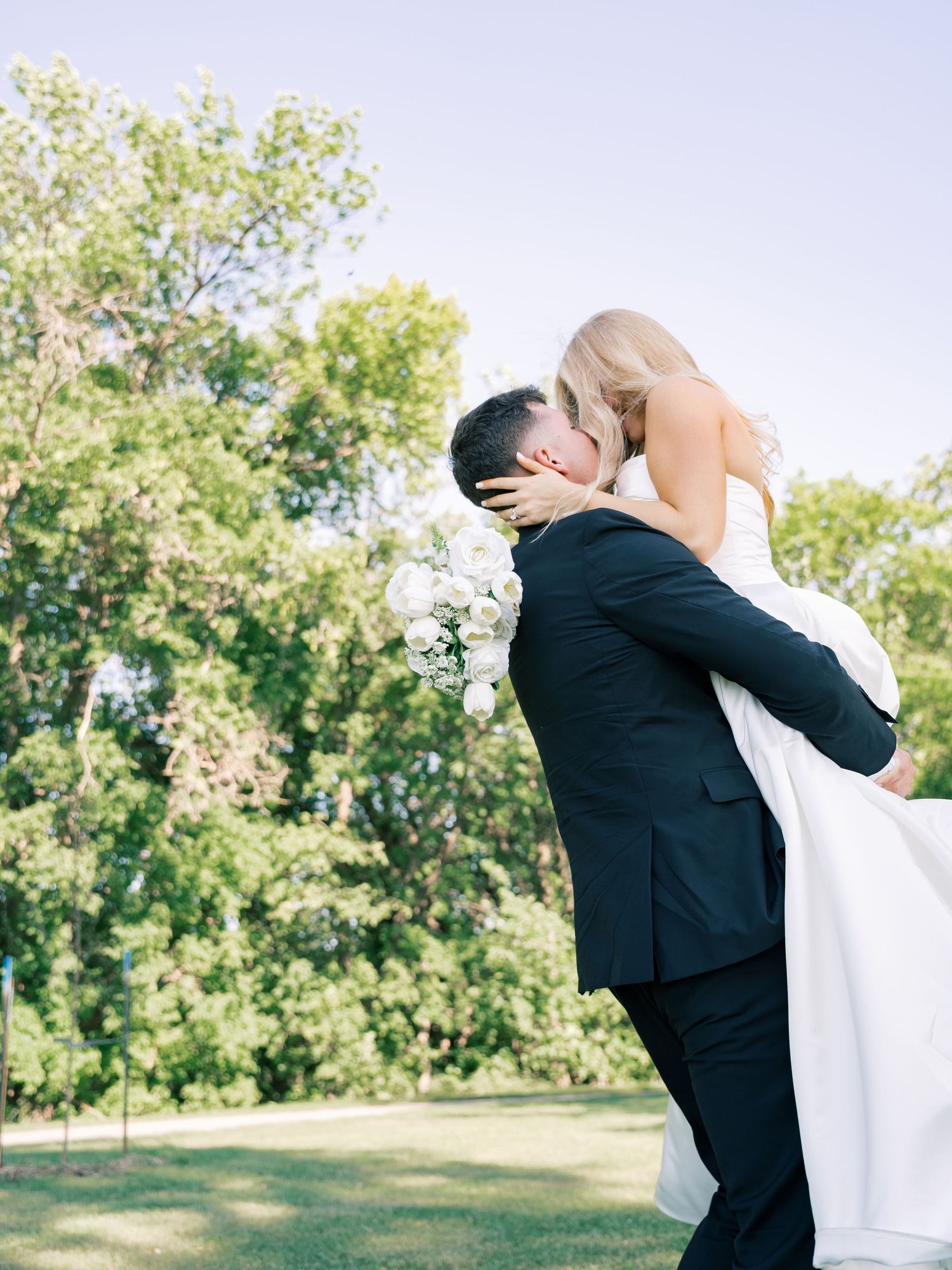 Groom in black suit lifts bride in white gown for a kiss outdoors, surrounded by lush green trees on their wedding day.