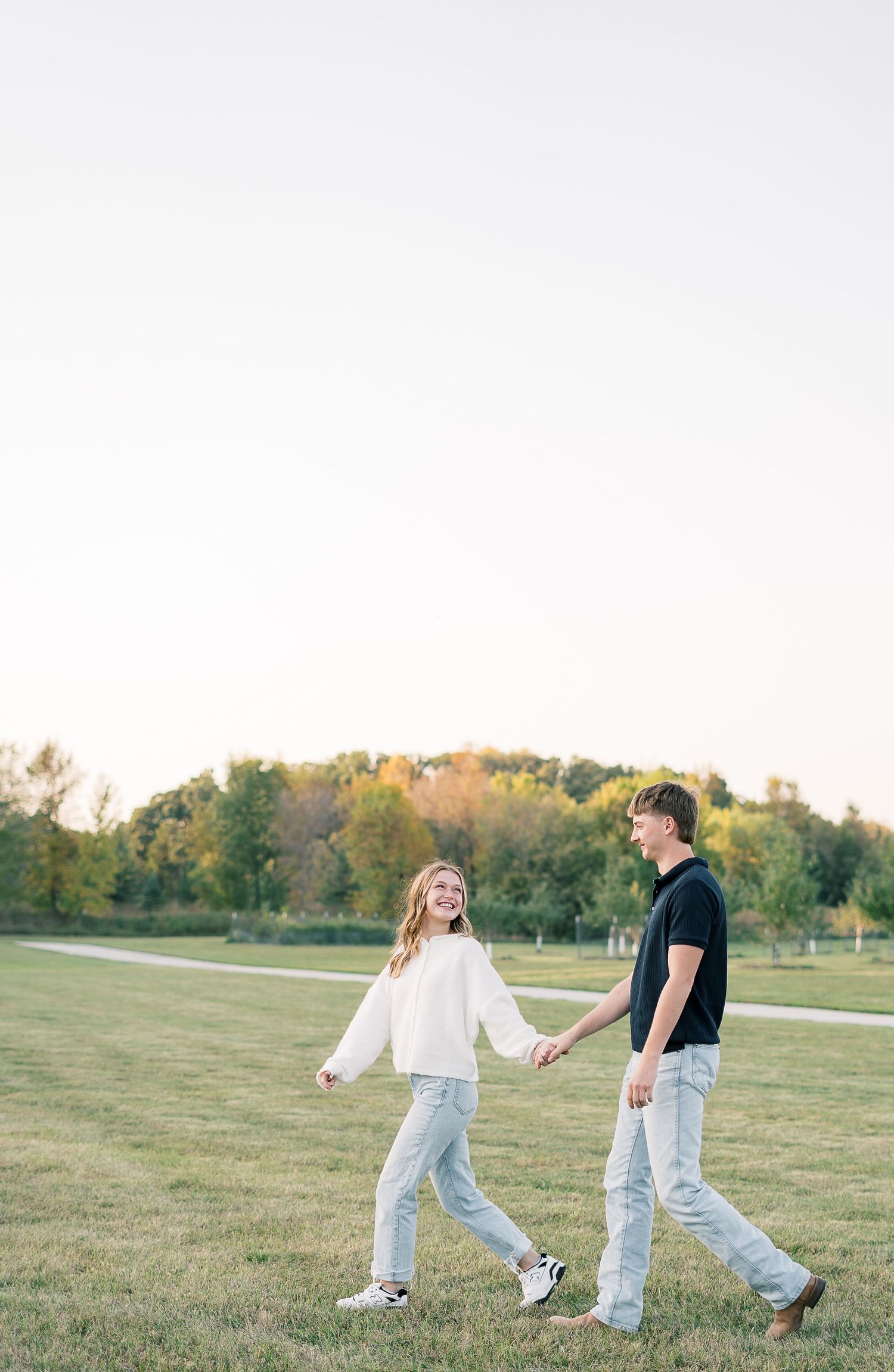 A couple walks playfully through a grassy field at sunset holding hands and dancing together.