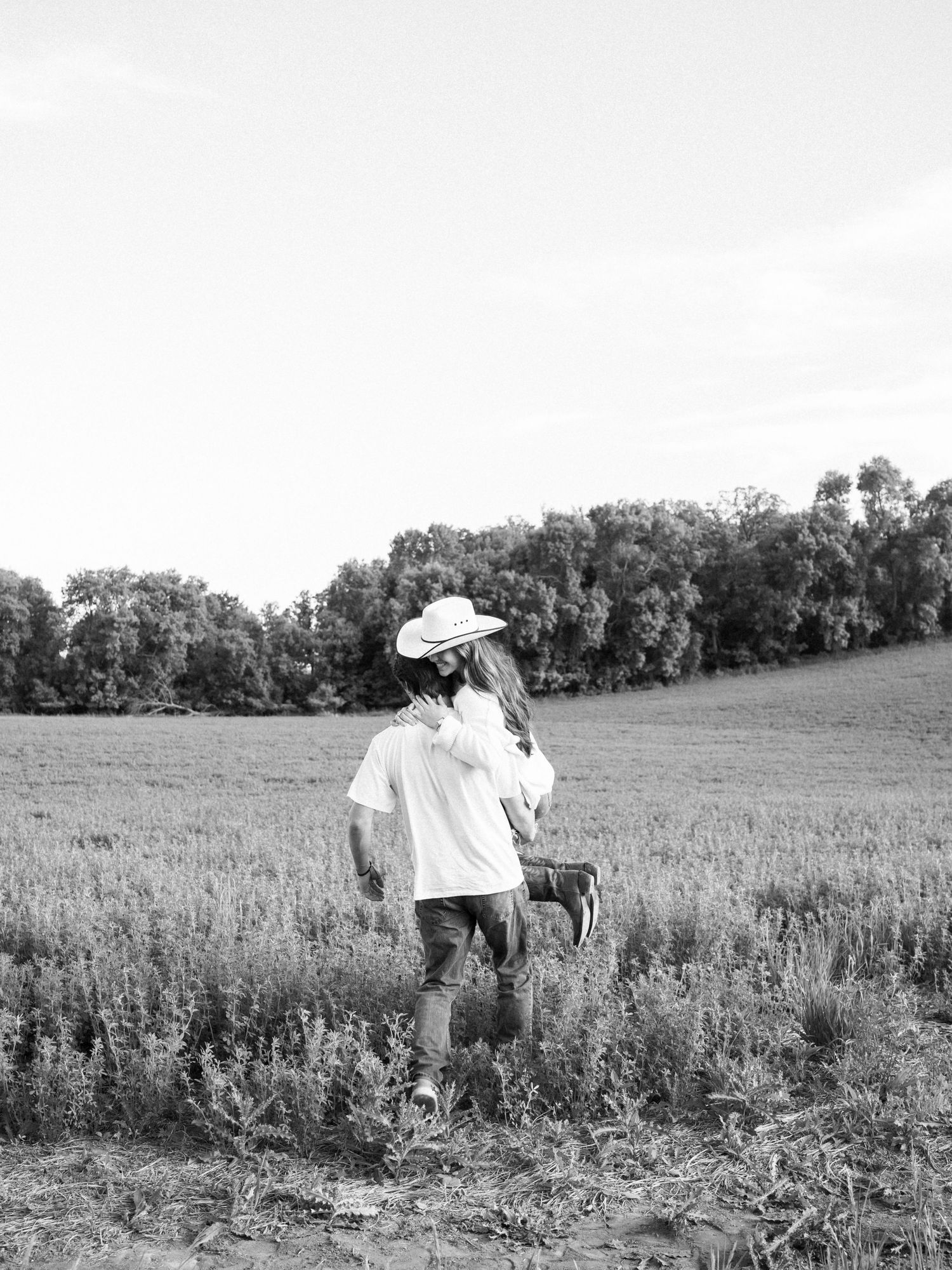 Black and white photo of a couple embracing in a lush green field, woman wearing a cowboy hat.