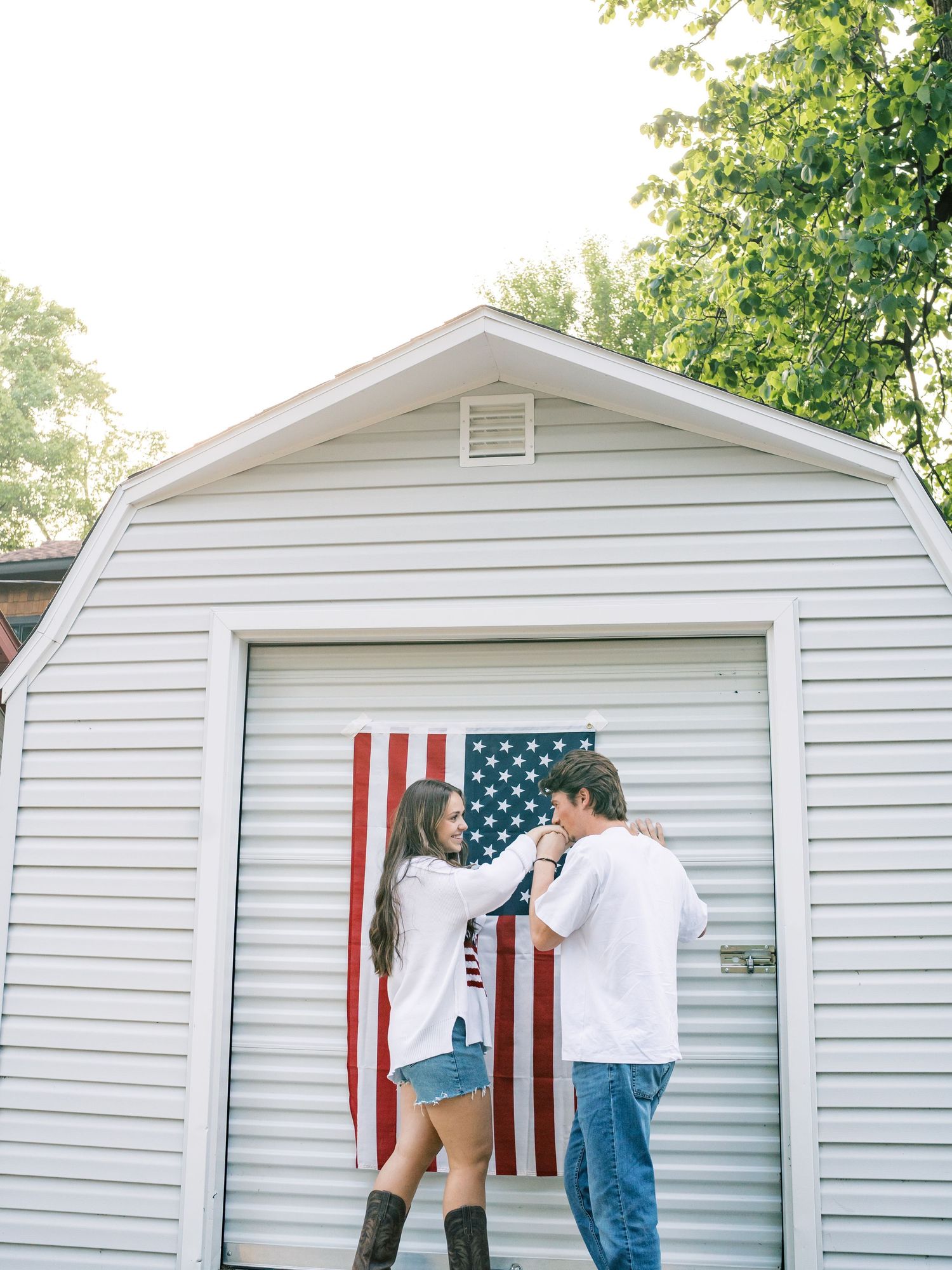 Couple dancing in front of a white garage with an American flag, wearing casual country attire during an engagement shoot.