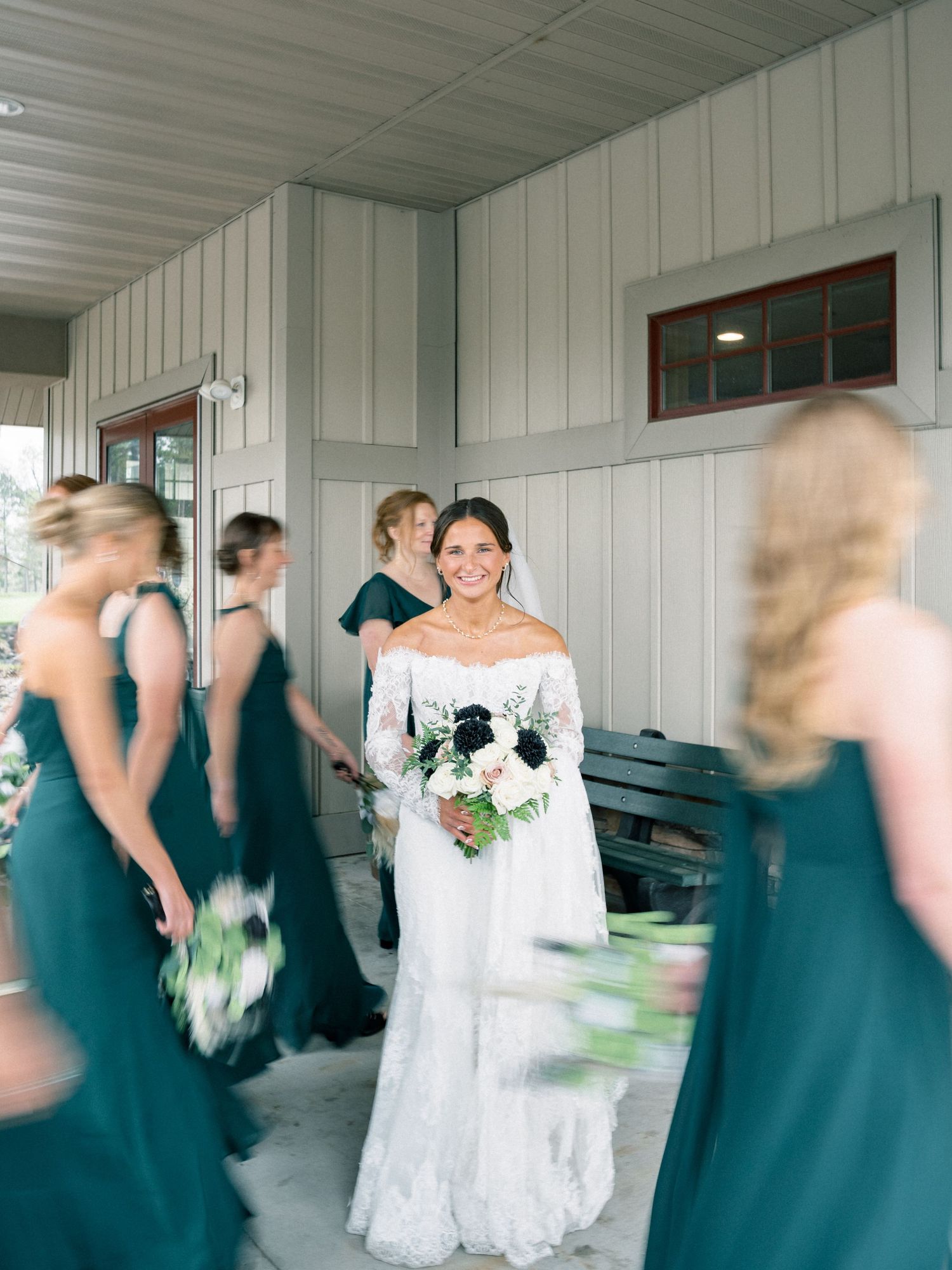 Bride in white lace gown holds bouquet surrounded by bridesmaids in teal dresses outside a rustic venue.