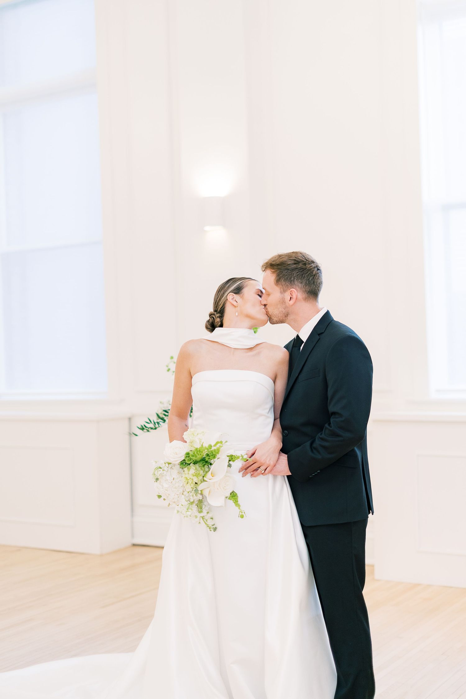 A romantic wedding couple shares a tender kiss in an elegant white room with hardwood floors and natural light.