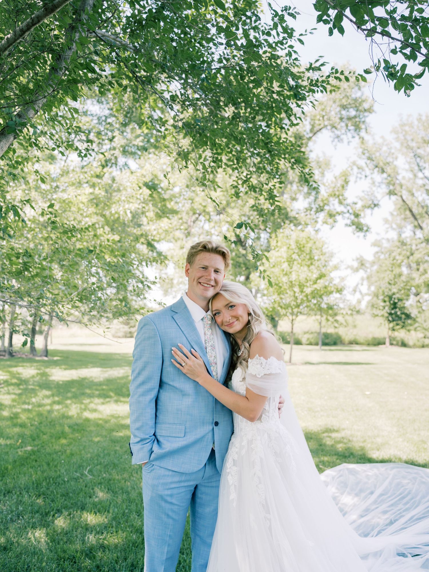 A couple poses outdoors under green trees, groom in light blue suit, bride in white wedding gown on a sunny day.