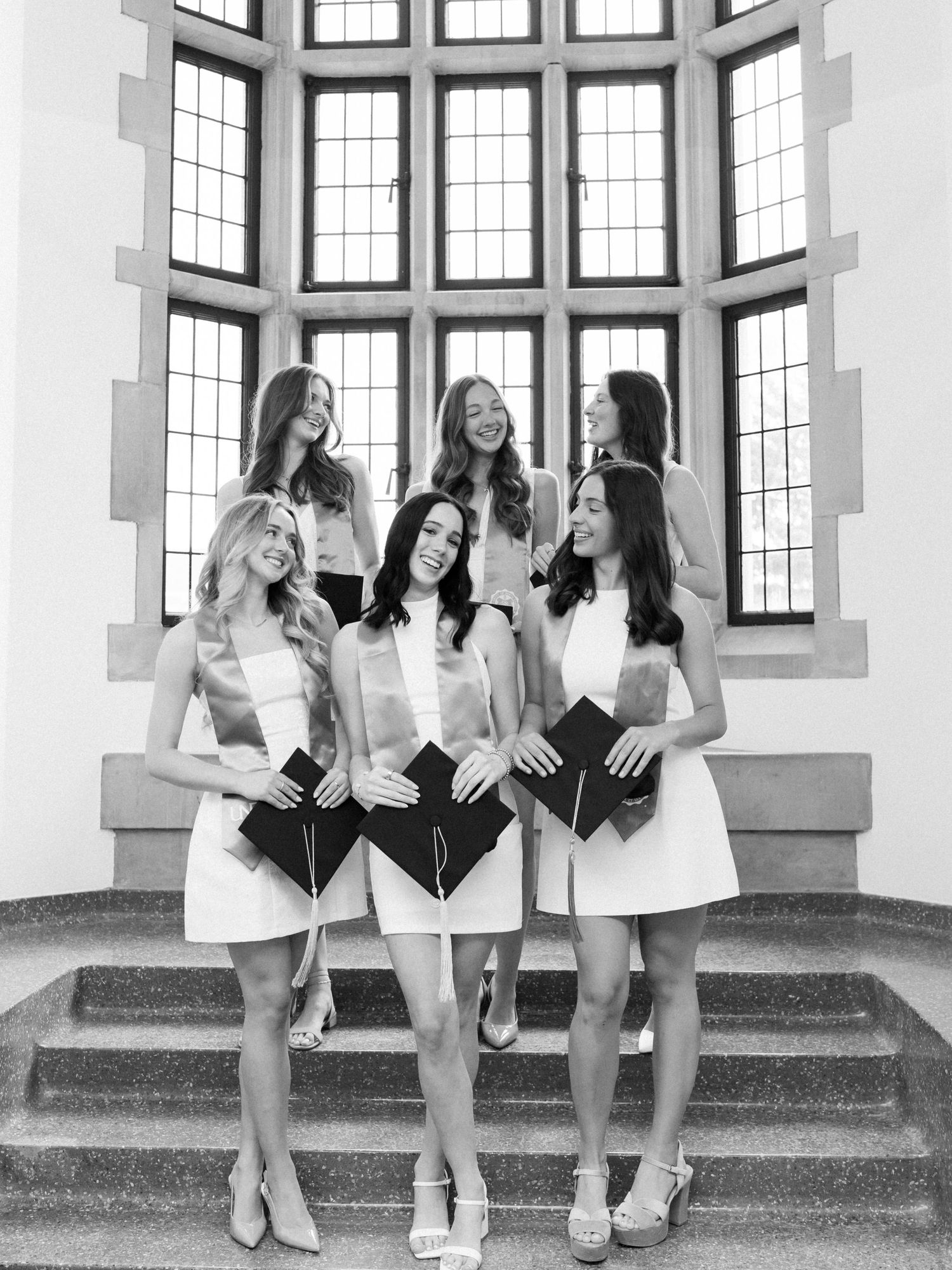 Six young women in white dresses holding graduation caps pose on stone steps in a black and white photo.