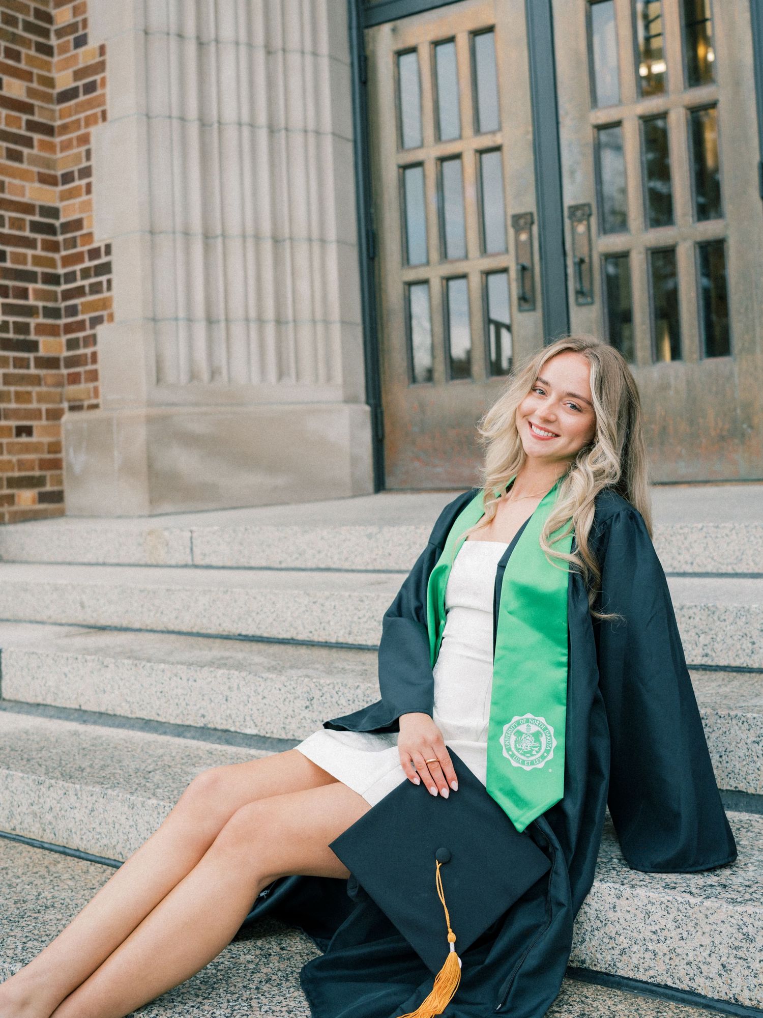 Smiling female graduate in dark cap and gown with green stole sits on university steps holding mortarboard.